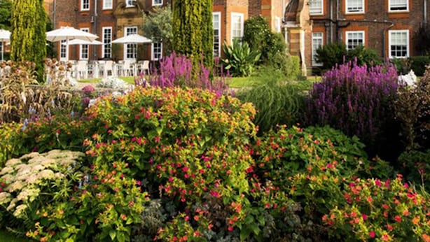 Un edificio grande e histórico con varias chimeneas, rodeado de un jardín bien cuidado con diversas plantas en flor.