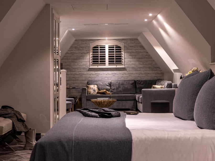 Um quarto de sótão aconchegante com teto inclinado, uma janela grande, um sofá cinza e uma cama com roupa de cama cinza.