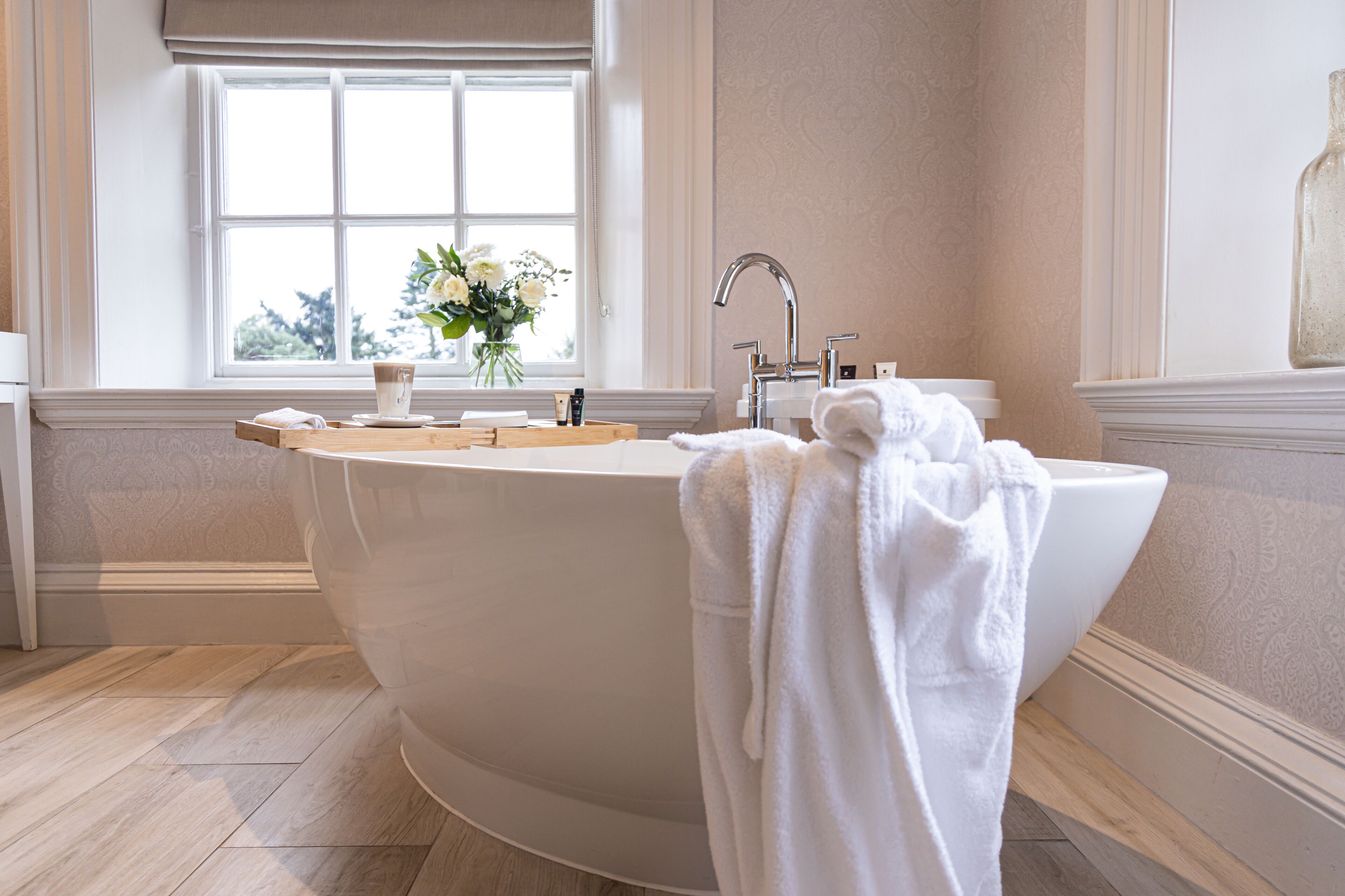 A large freestanding bathtub in a bathroom with a window, a vase of flowers, and a towel on the side.