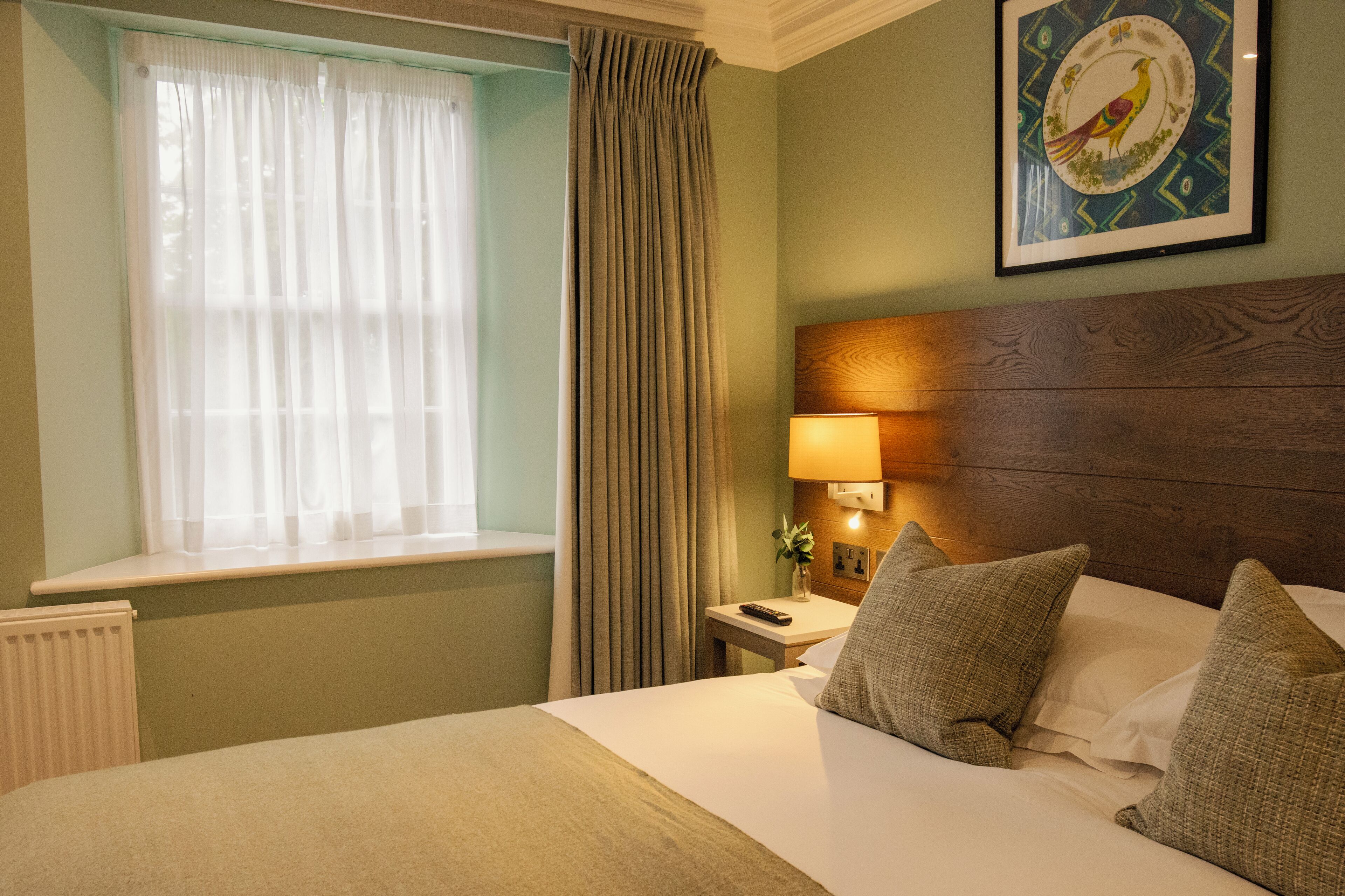 A hotel room with a bed, a nightstand, a lamp, a window with curtains, and a wall-mounted picture.