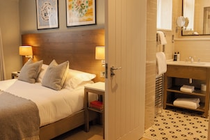 Habitación de hotel con cama, lámparas de noche, mesita de noche, cabecera de madera y baño con lavamanos, espejo y toallero.