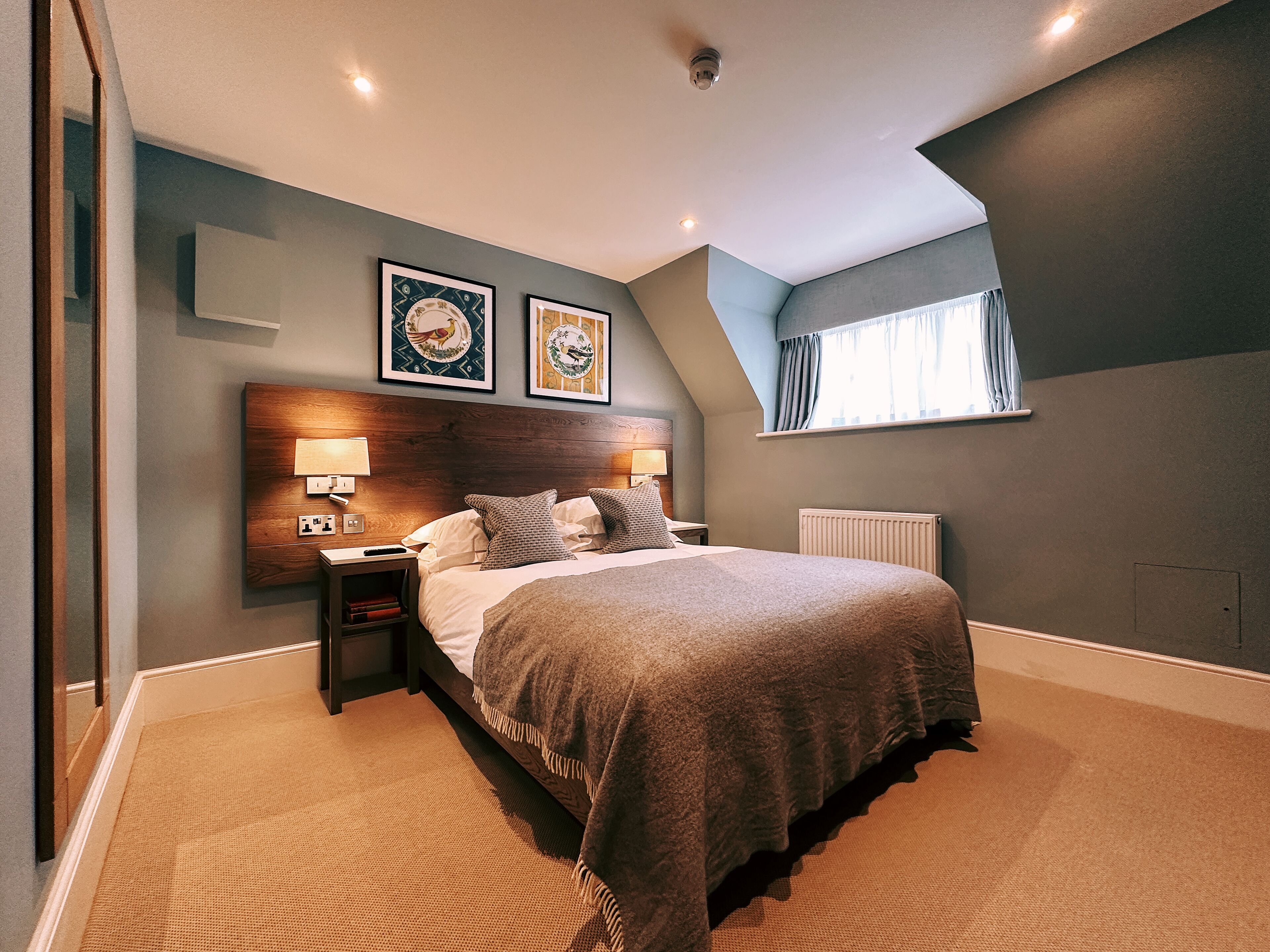 A modern bedroom with a large bed, a wooden headboard, two bedside tables with lamps, and two framed pictures on the wall.
