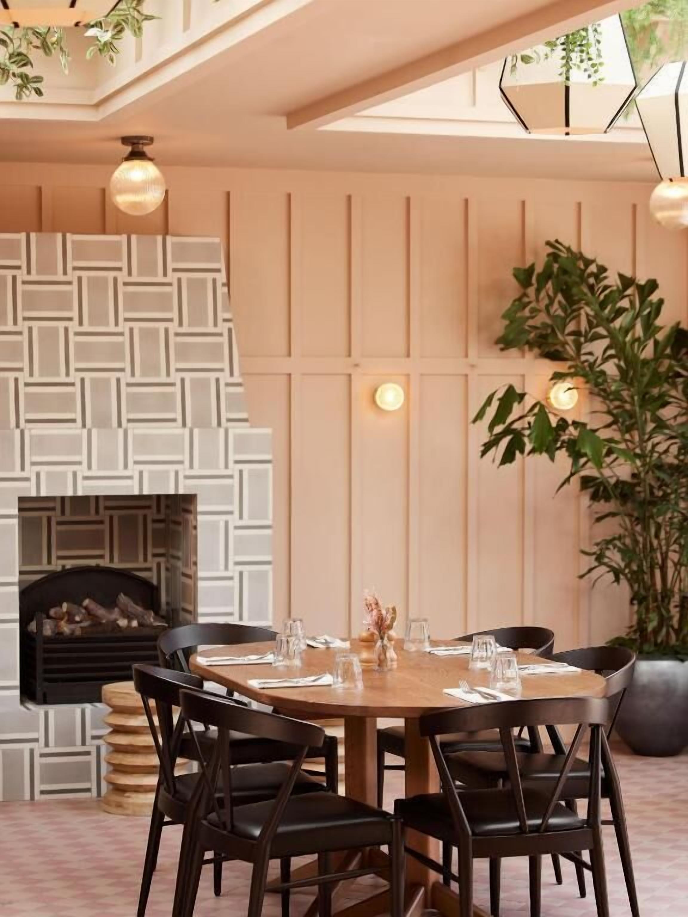A dining area with a round table, black chairs, a geometric wall pattern, a fireplace, and hanging plants.