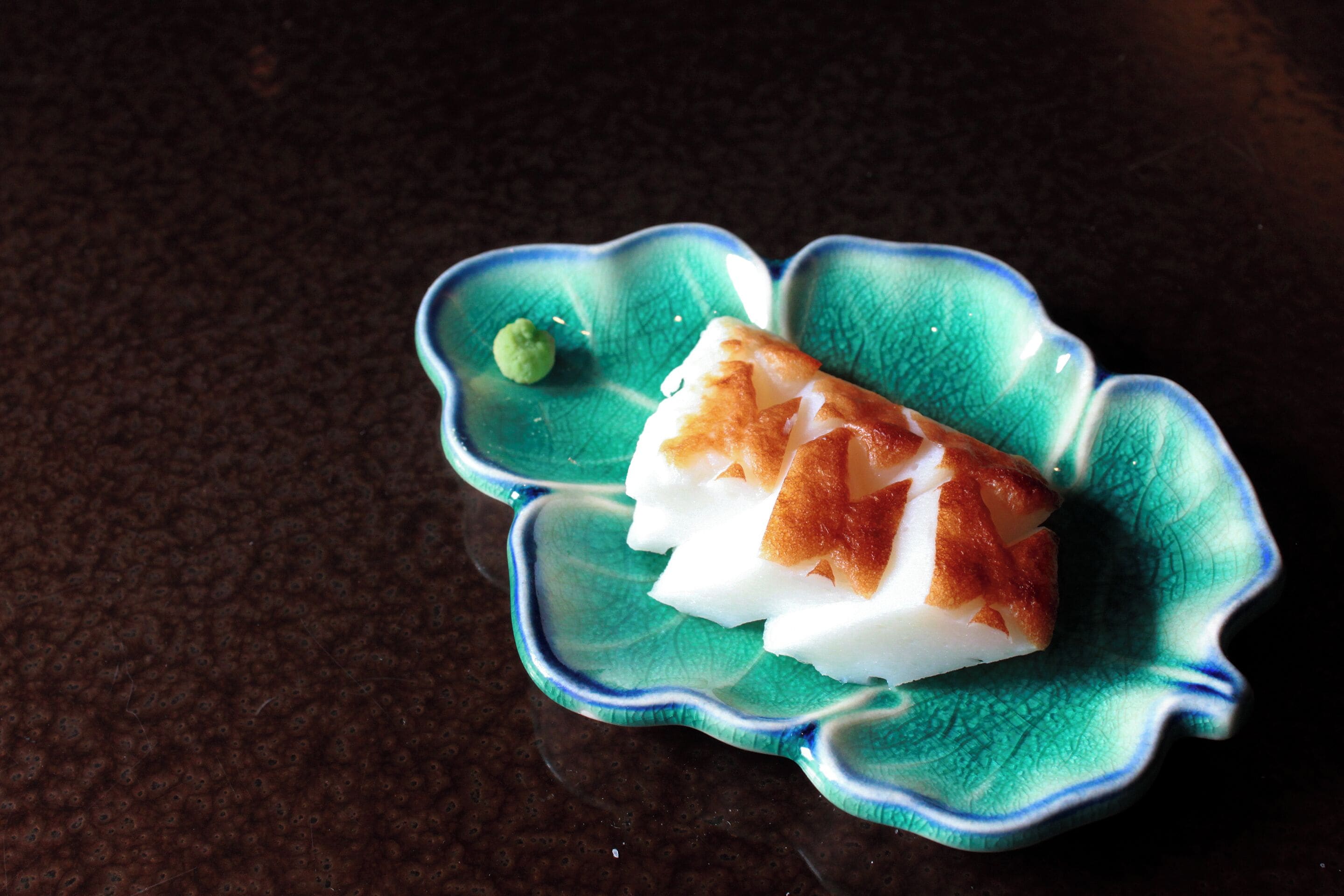 A green ceramic dish with a scalloped edge containing a white and brown food item, possibly a fish or meat, and a small green garnish.