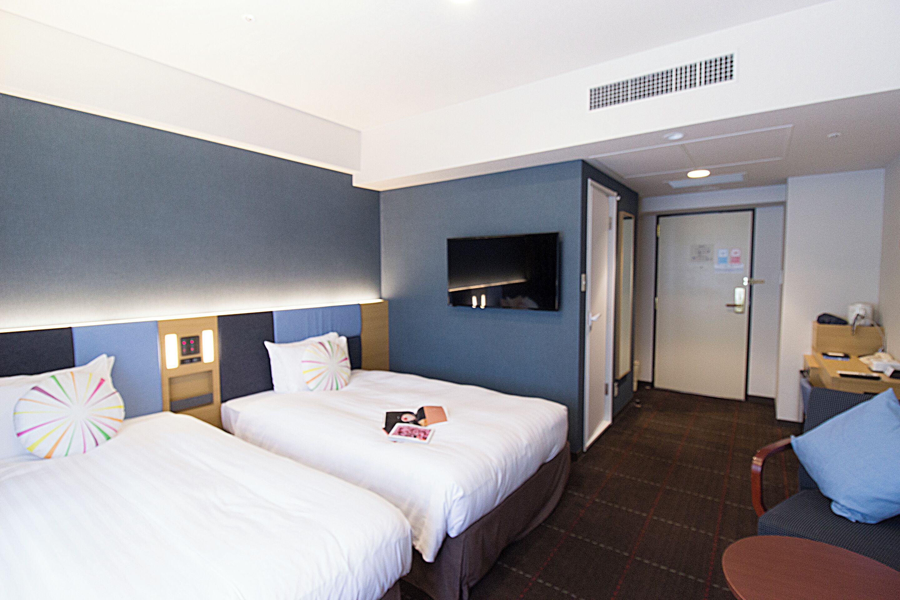 Quarto de hotel com duas camas, televisão na parede, escrivaninha com cadeira e porta para outro cômodo.