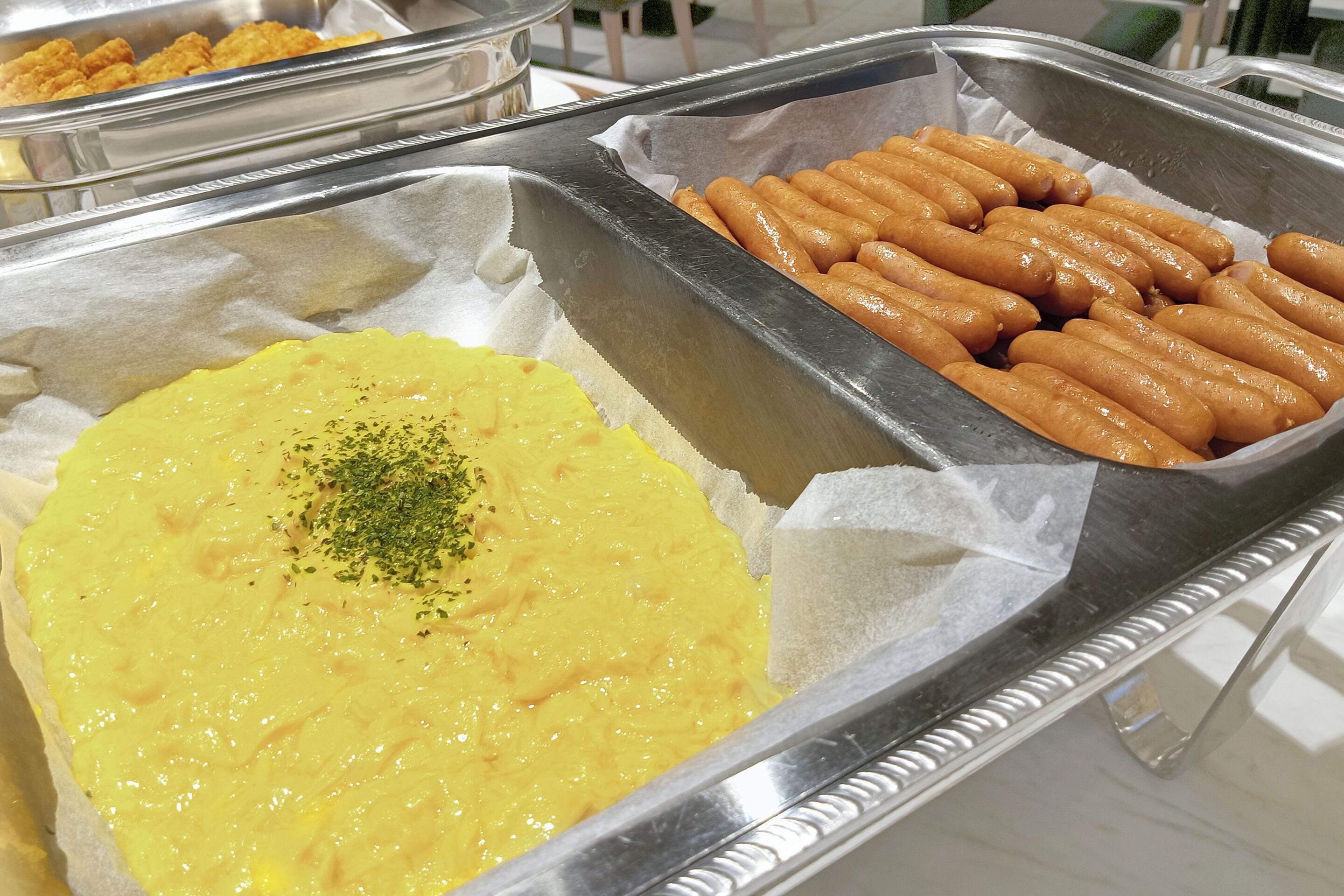 Een buffet met diverse gerechten, waaronder een grote portie gele dipsaus, meerdere hotdogs en verschillende gebakken snacks.
