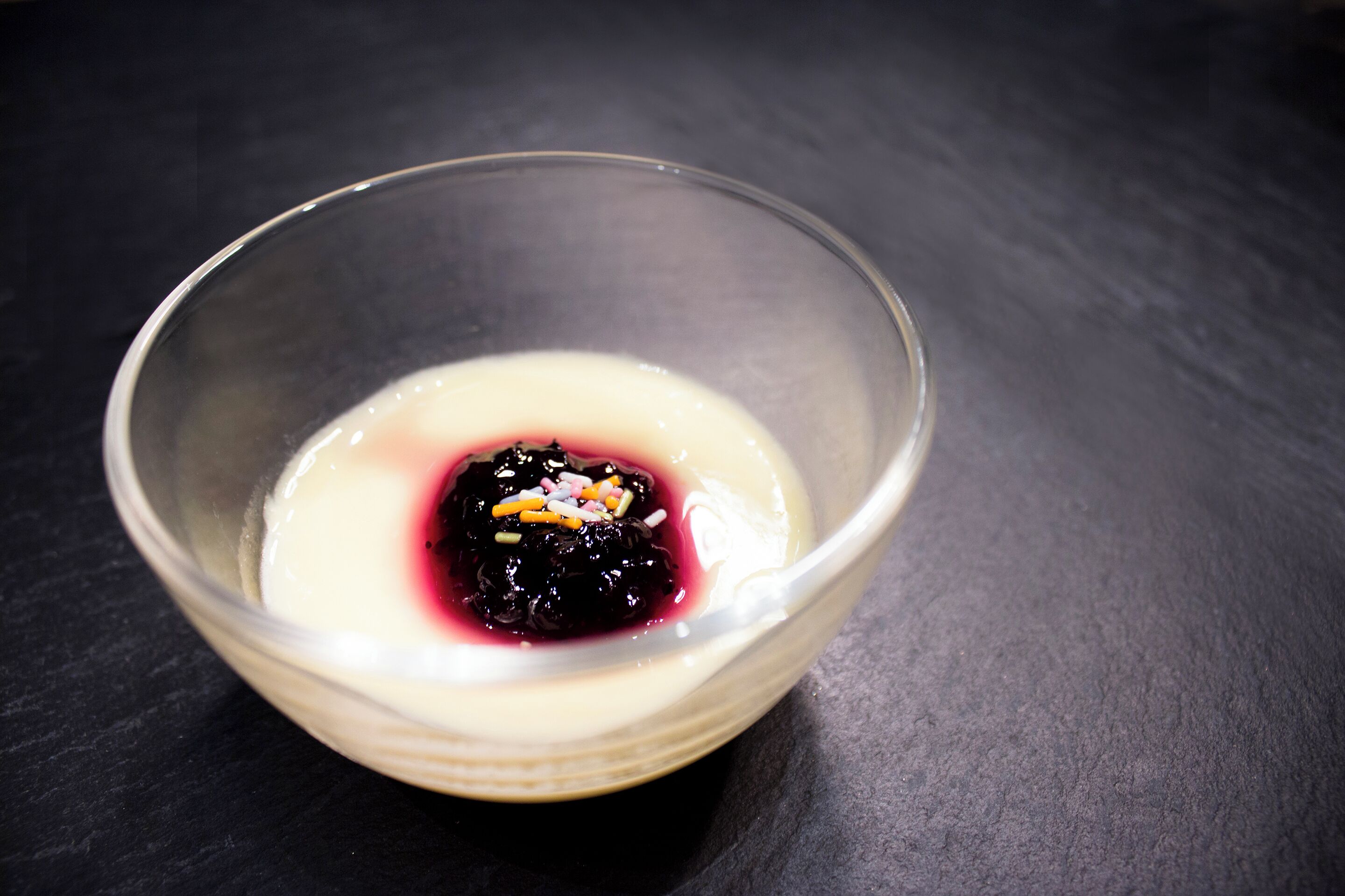 Een glazen schaal met een dessert, met een donkere topping en een romige basis, tegen een donkere achtergrond.