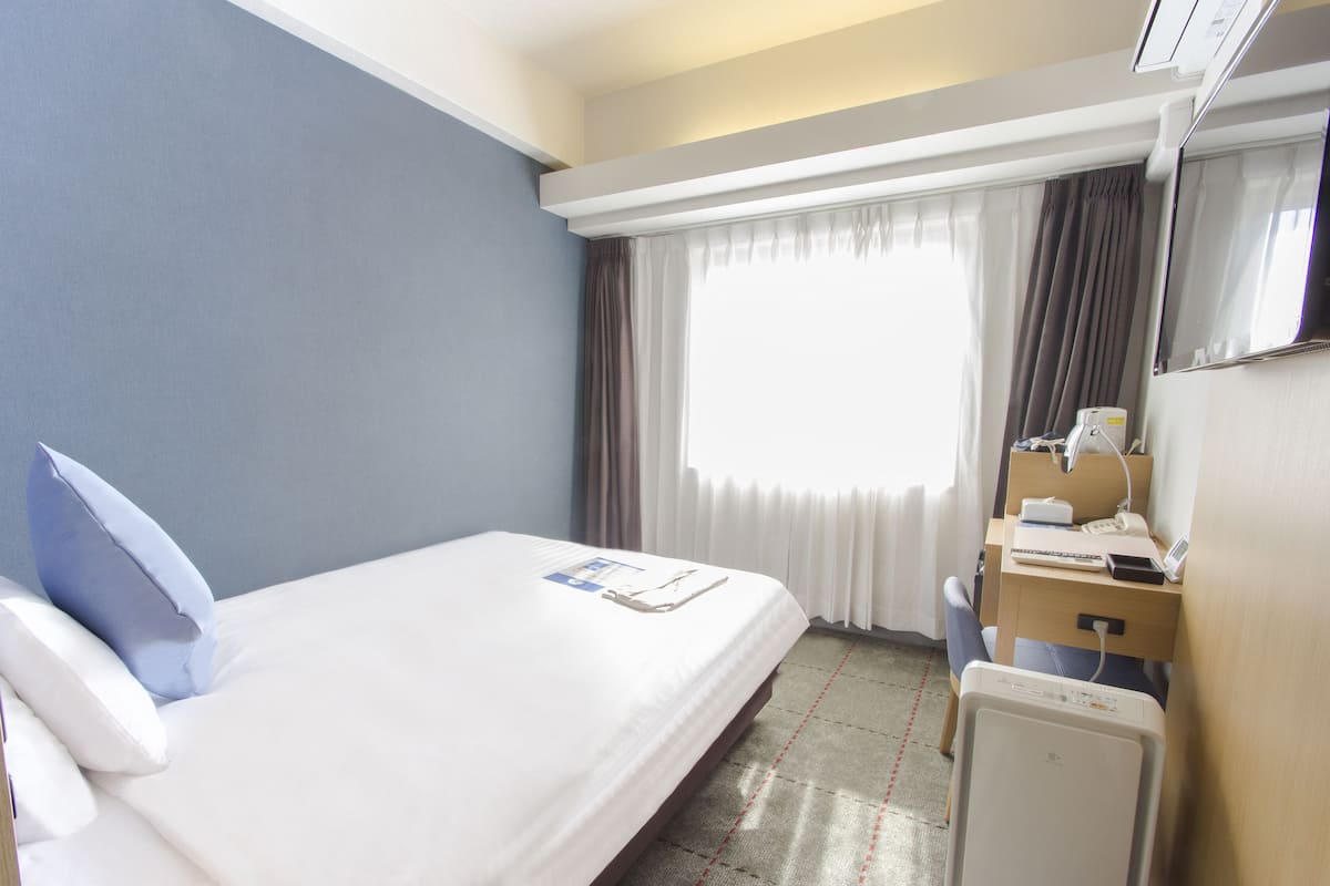 Une chambre d’hôtel avec un lit, un bureau, une chaise, une télévision et une fenêtre avec des rideaux.