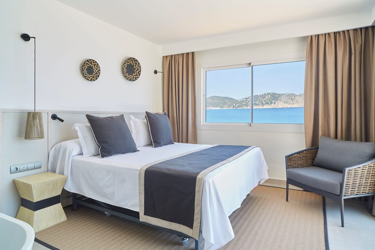 A hotel room with a bed, a chair, a view of the sea, and a window with curtains.