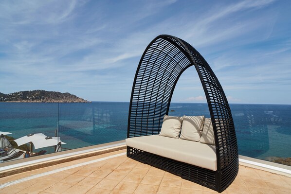 A curved black wicker sofa with beige cushions on a balcony overlooking a clear blue sea and a distant island. - Invisa Hotel Club Cala Verde (Santa Eulalia del Rio)