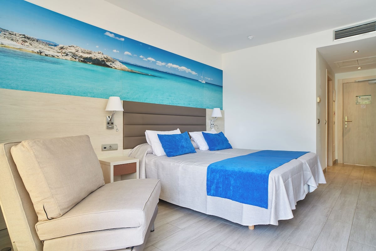 A hotel room with a bed, a chair, a nightstand, and a painting of a beach scene.