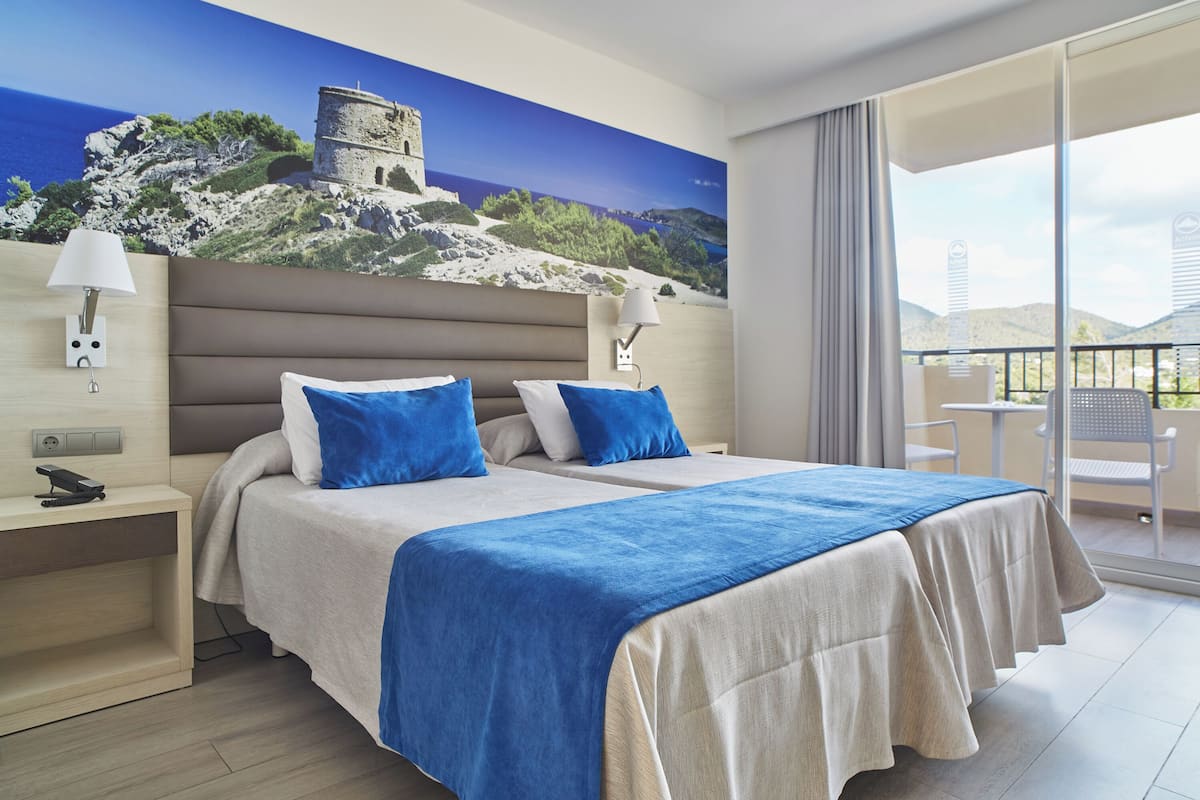 A hotel room with two beds, a balcony with a view of the sea and a castle, and a painting of the same scene on the wall.
