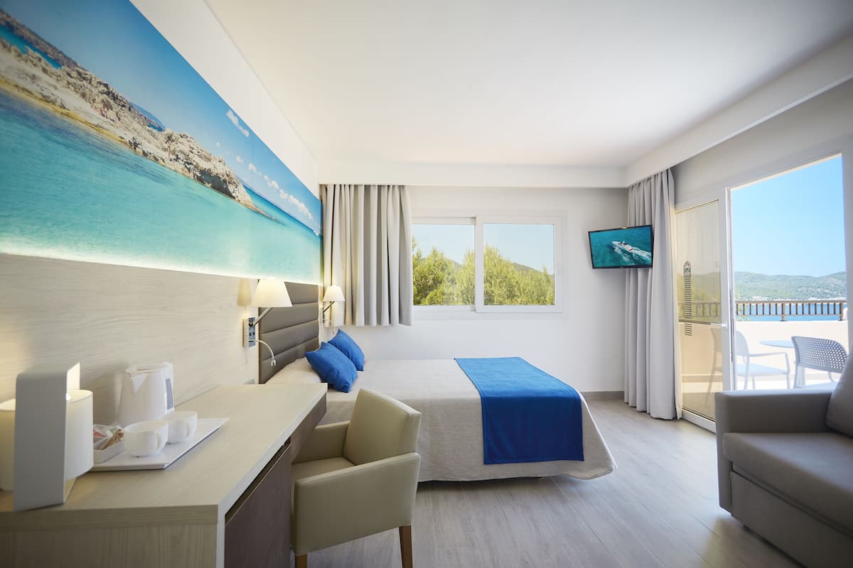 A hotel room with a bed, a desk with a laptop, a chair, a TV, and a balcony with a view of the sea.