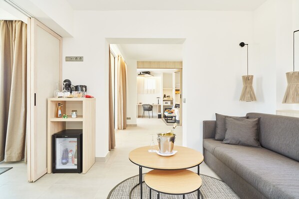 A modern living room with a sofa, a small table, a mini-fridge, and a dining area in the background.