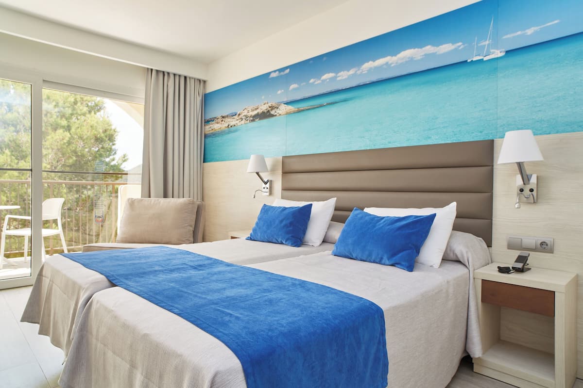 A hotel room with two beds, a balcony, a painting of a beach, and a nightstand with a lamp.
