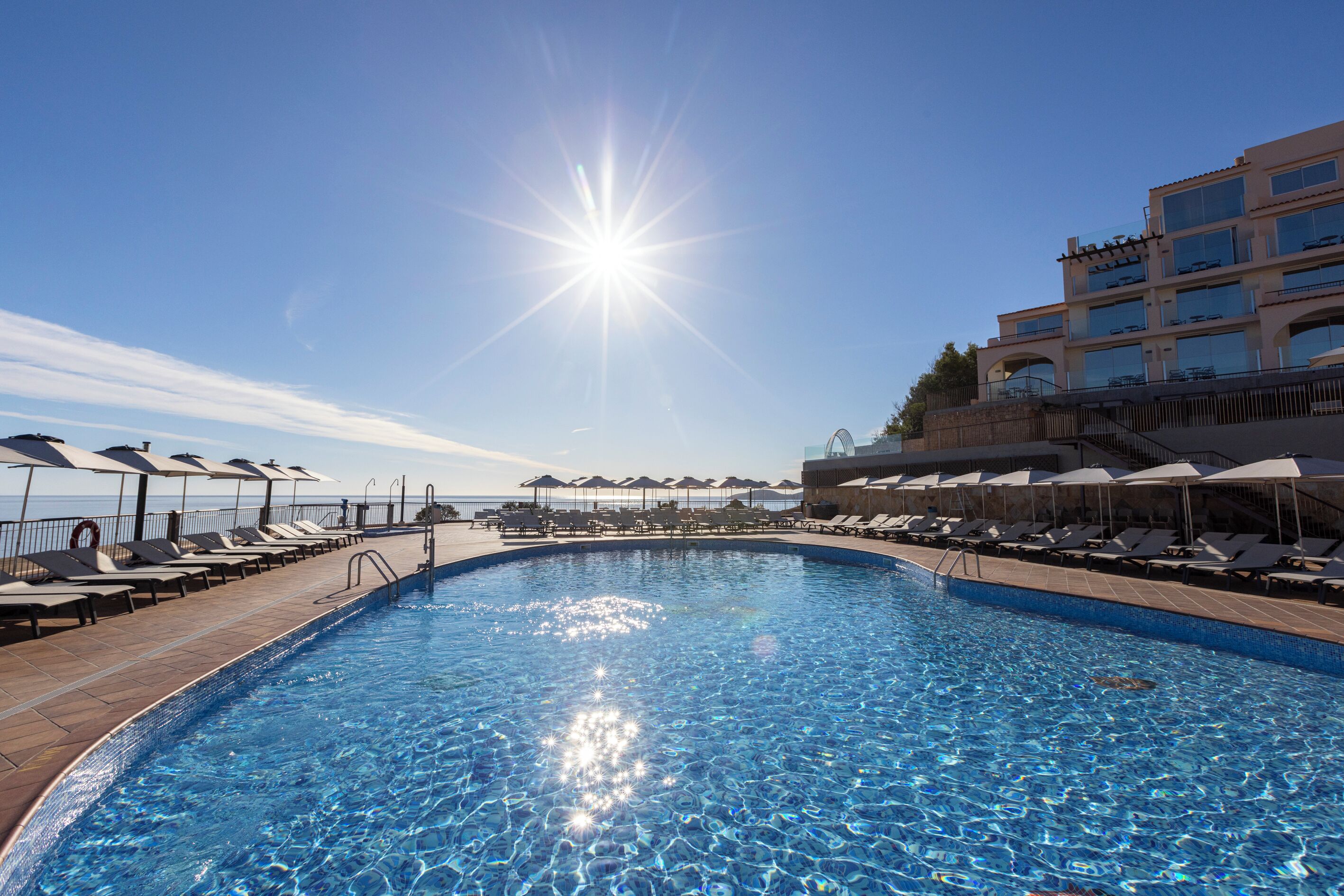 Una piscina con reflejos de sol en el agua, rodeada de sillones de playa y sombrillas, con un edificio al fondo.