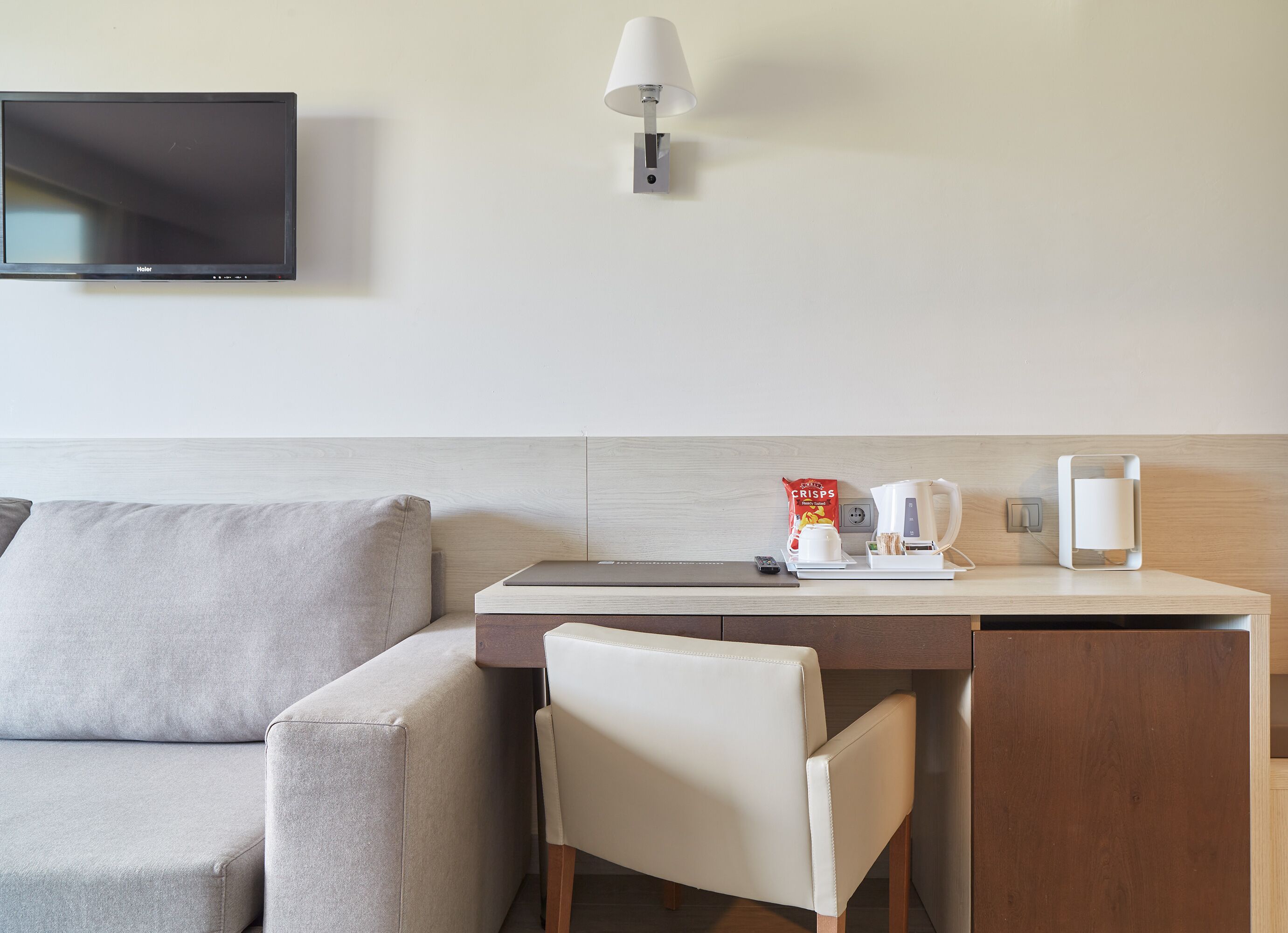 Una habitación de hotel moderna con televisión de pantalla plana montada en la pared, una pequeña mesa con cafetera, una bolsa de patatas fritas y un control remoto.