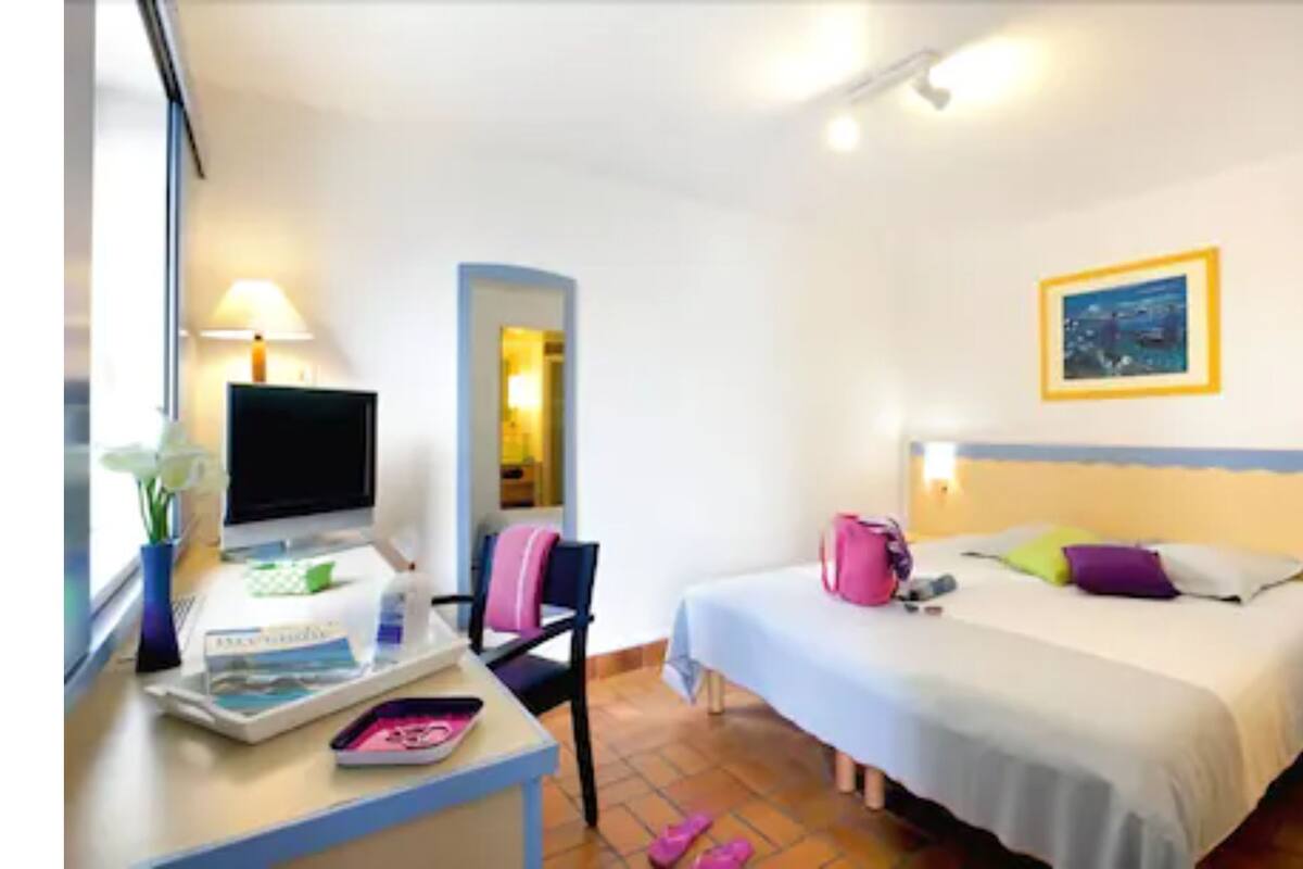 A hotel room with a bed, bedside table, lamp, TV, and a window with curtains.