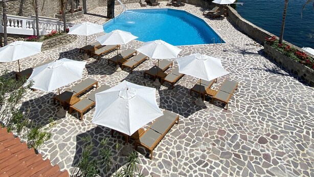 Un área junto a la piscina con sillones de descanso y sombrillas, un suelo de baldosas de piedra y vista al mar.