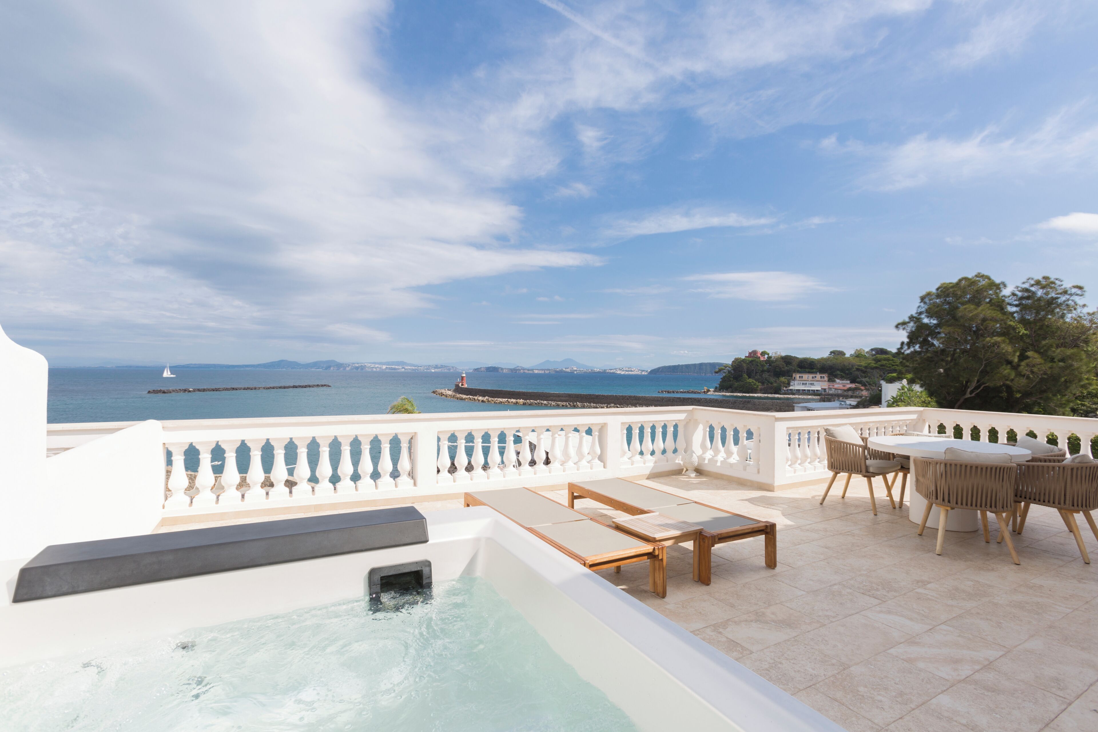 Une terrasse sur le toit dotée d’un jacuzzi, de chaises longues et d’une table, offrant une vue imprenable sur la côte, avec une jetée et des montagnes lointaines.