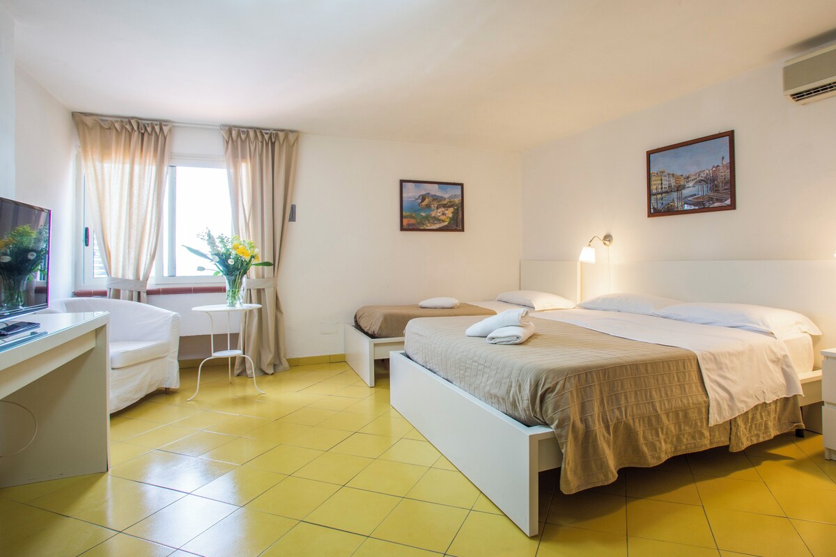 Une chambre d’hôtel avec un lit, un bureau, une chaise, une petite table avec un vase et deux tableaux accrochés au mur.