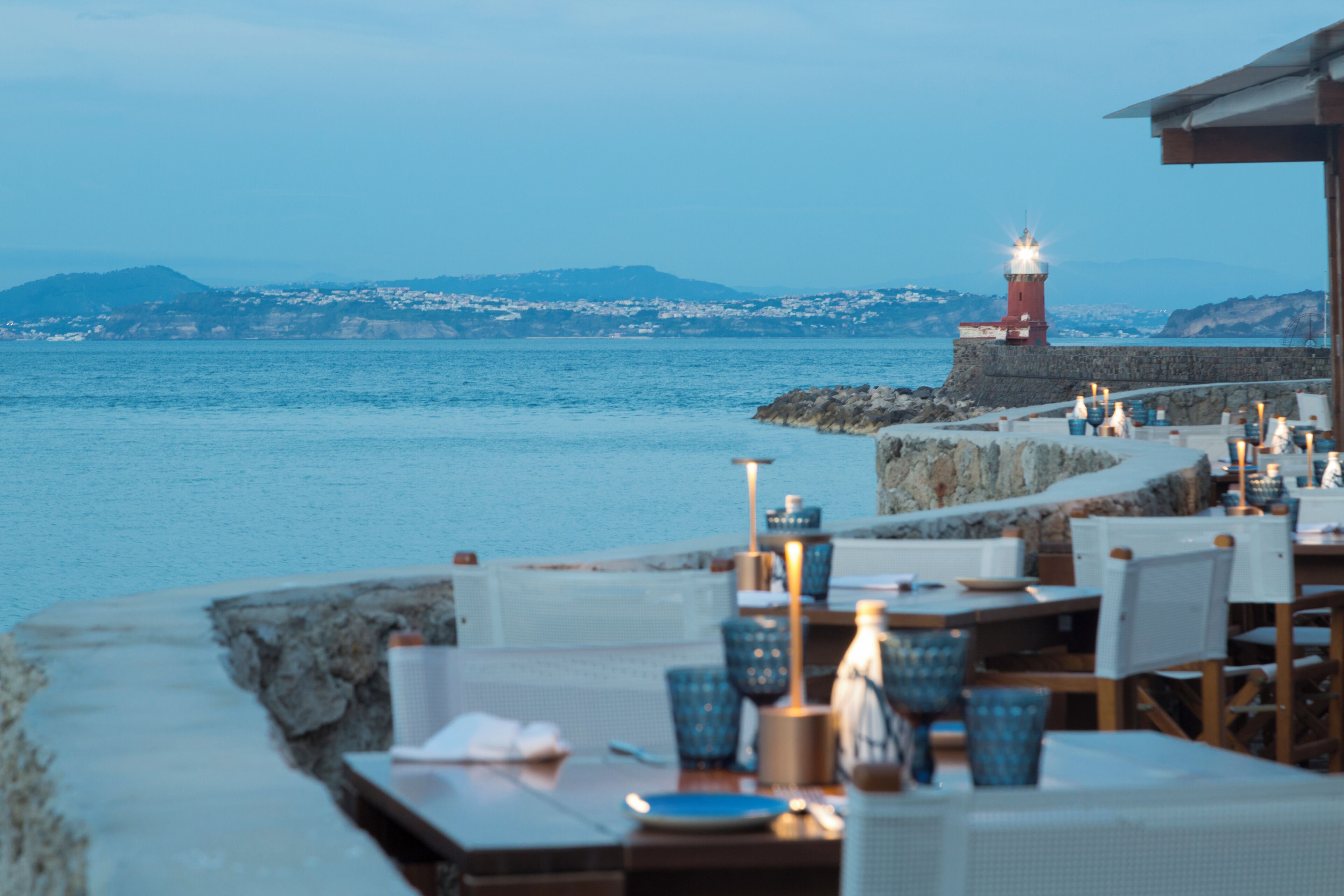 A seaside restaurant with tables set for dining, a lighthouse in the background, and a rocky shoreline.