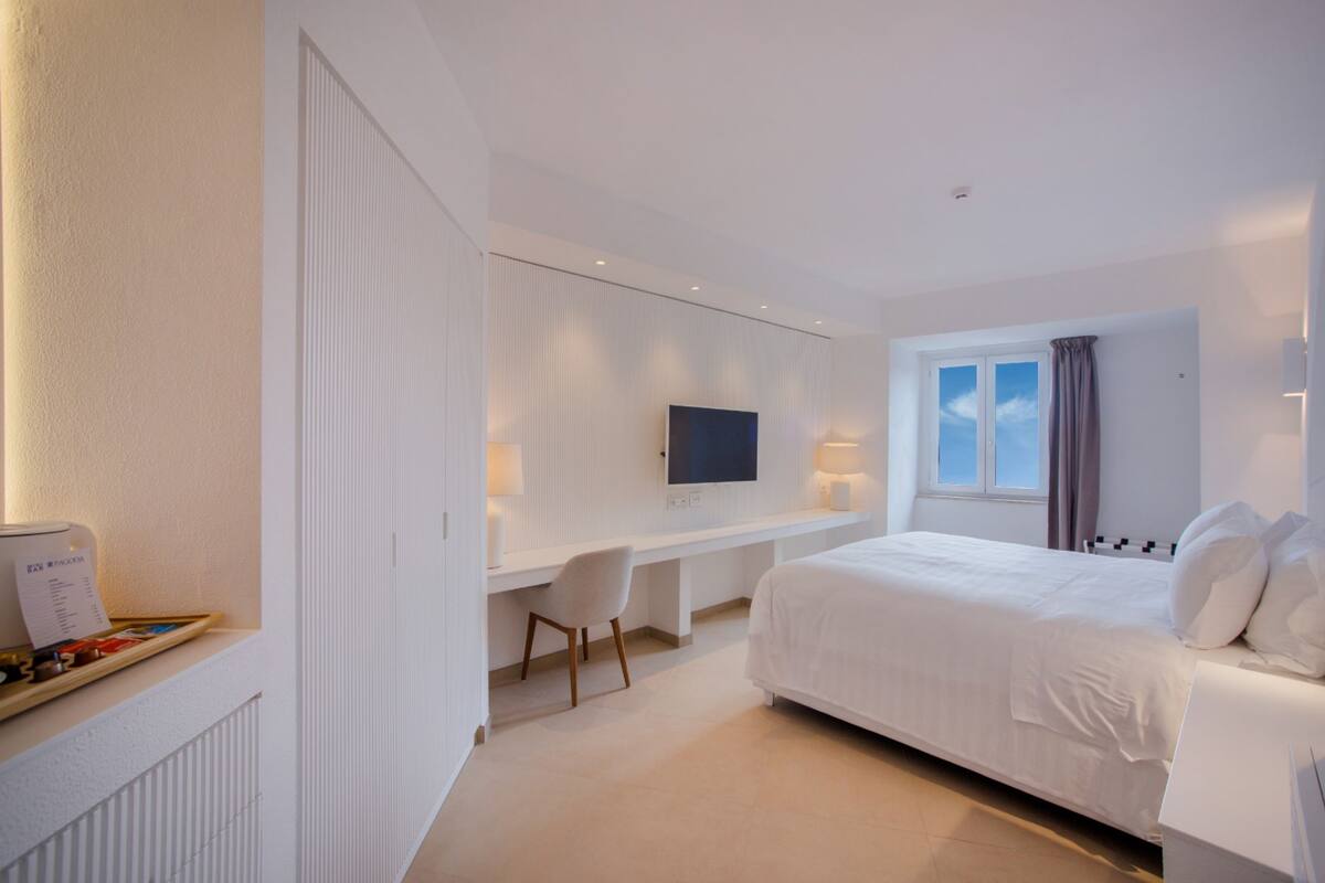 A modern hotel room with a large bed, a desk with a chair, a flat-screen TV, and a window with curtains.