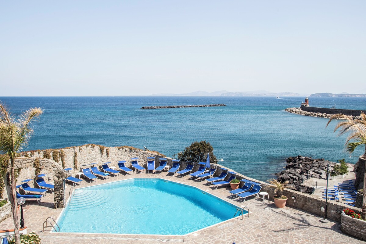 Una piscina con sillas reclinables azules, vista despejada al mar y un rompeolas rocoso.