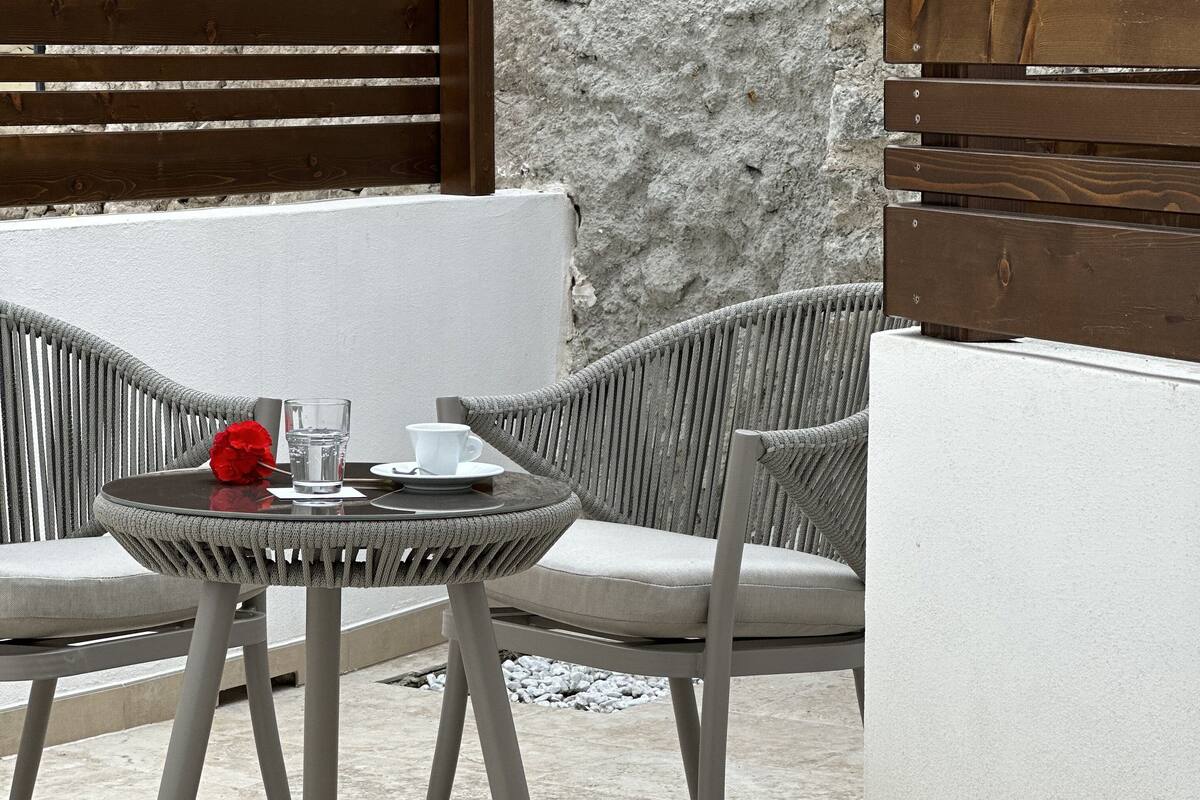A modern outdoor seating area with a round table and two grey wicker chairs, a white cushioned sofa, and a stone wall backdrop.