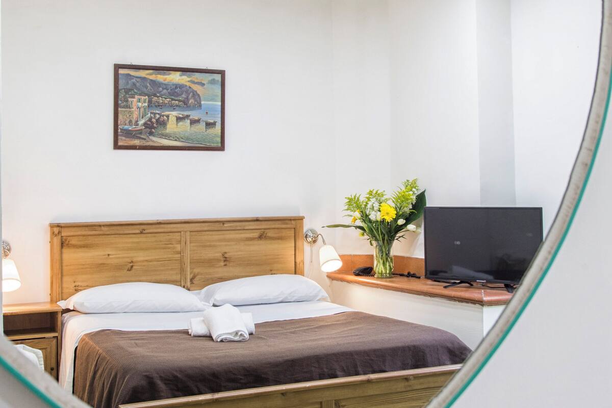A bedroom with a bed, bedside table, lamp, TV, and a painting on the wall.