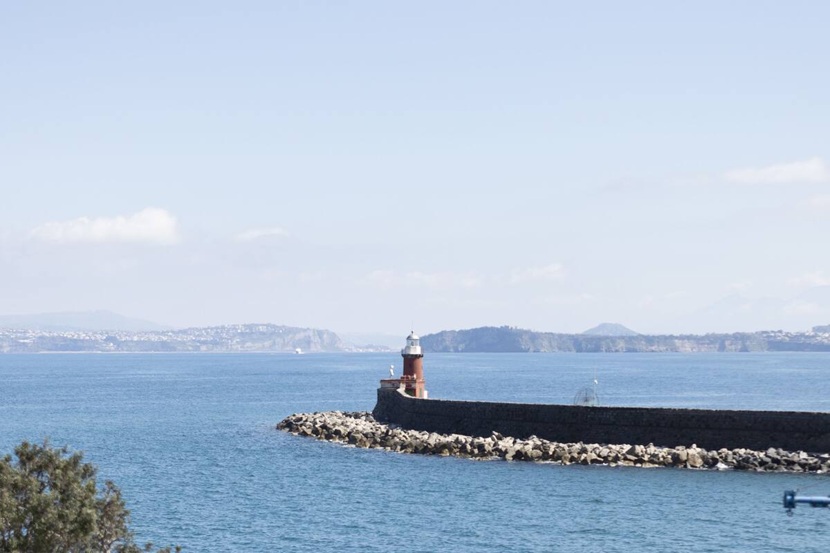 A lighthouse on a rocky pier extending into a calm sea, with distant hills and a clear sky.