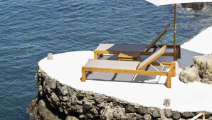 Un espacio de descanso junto al mar con una sombrilla blanca y sillones de madera sobre una plataforma rocosa.