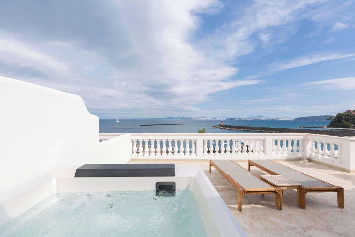 A rooftop terrace with a hot tub, lounge chairs, and a view of the sea and mountains.