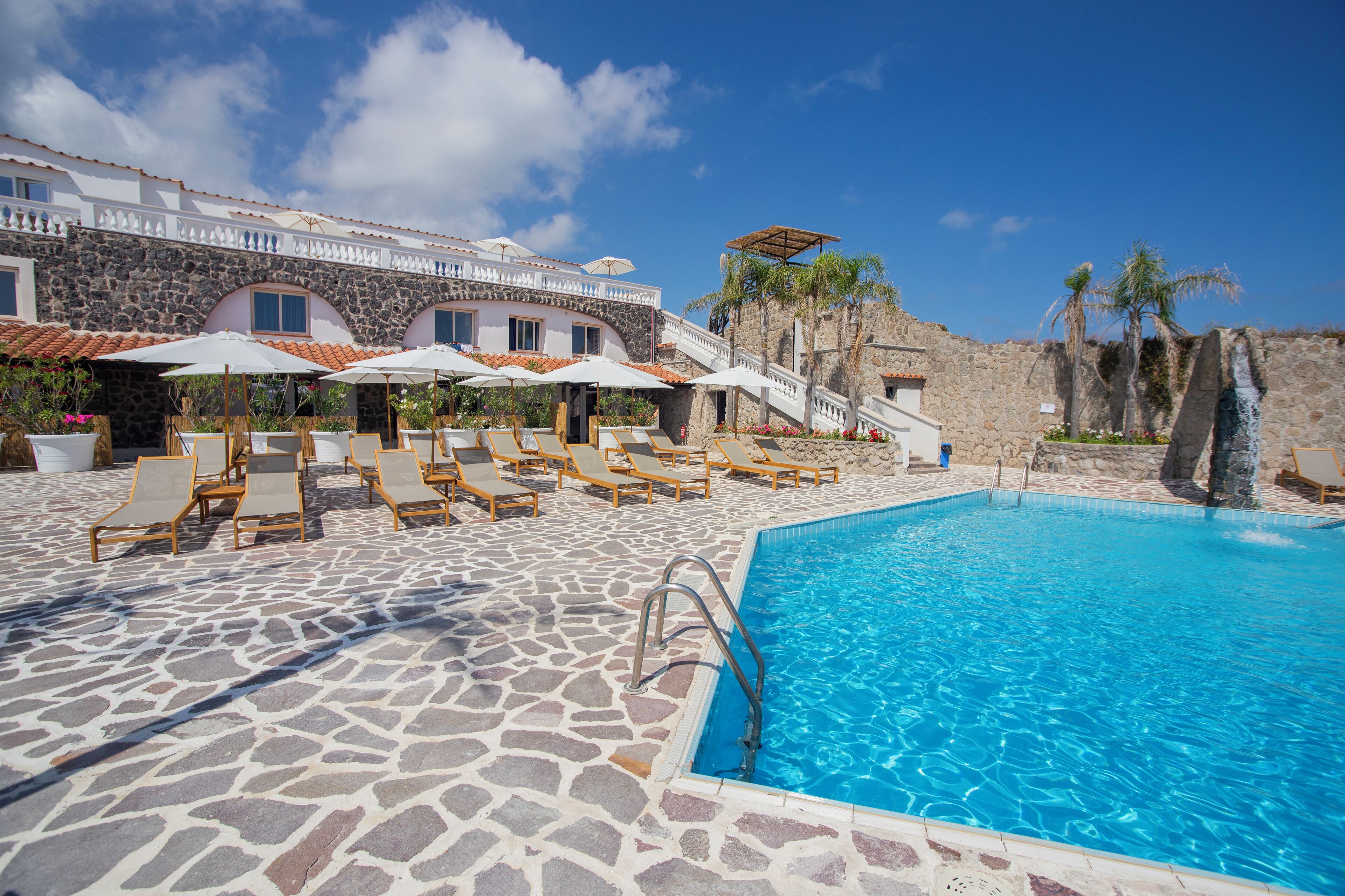A pool area with lounge chairs and umbrellas, a stone pool, and a building with a stone facade and arched windows.