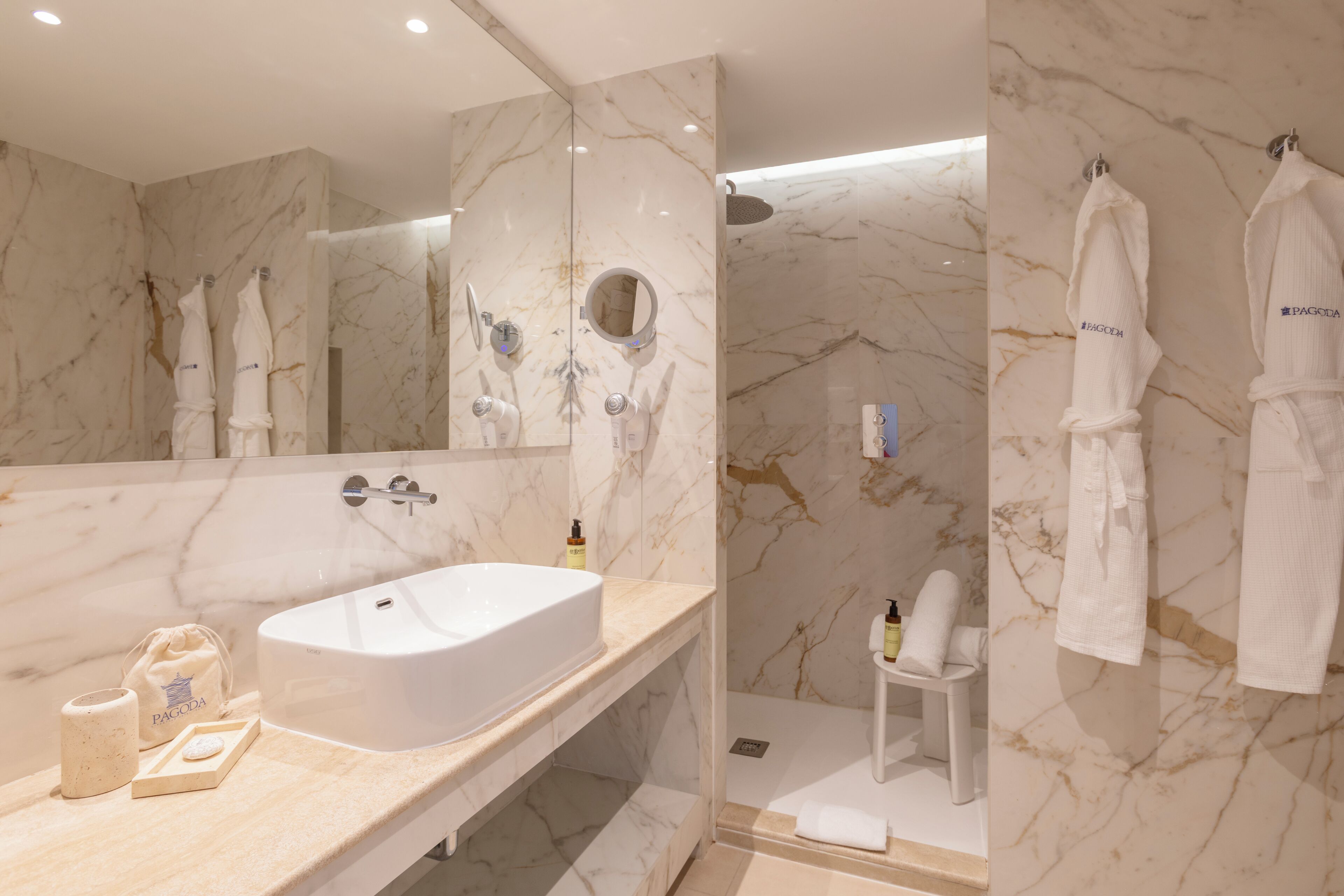 A modern bathroom with a marble sink, a large mirror, a shower area, and two white robes hanging on hooks.