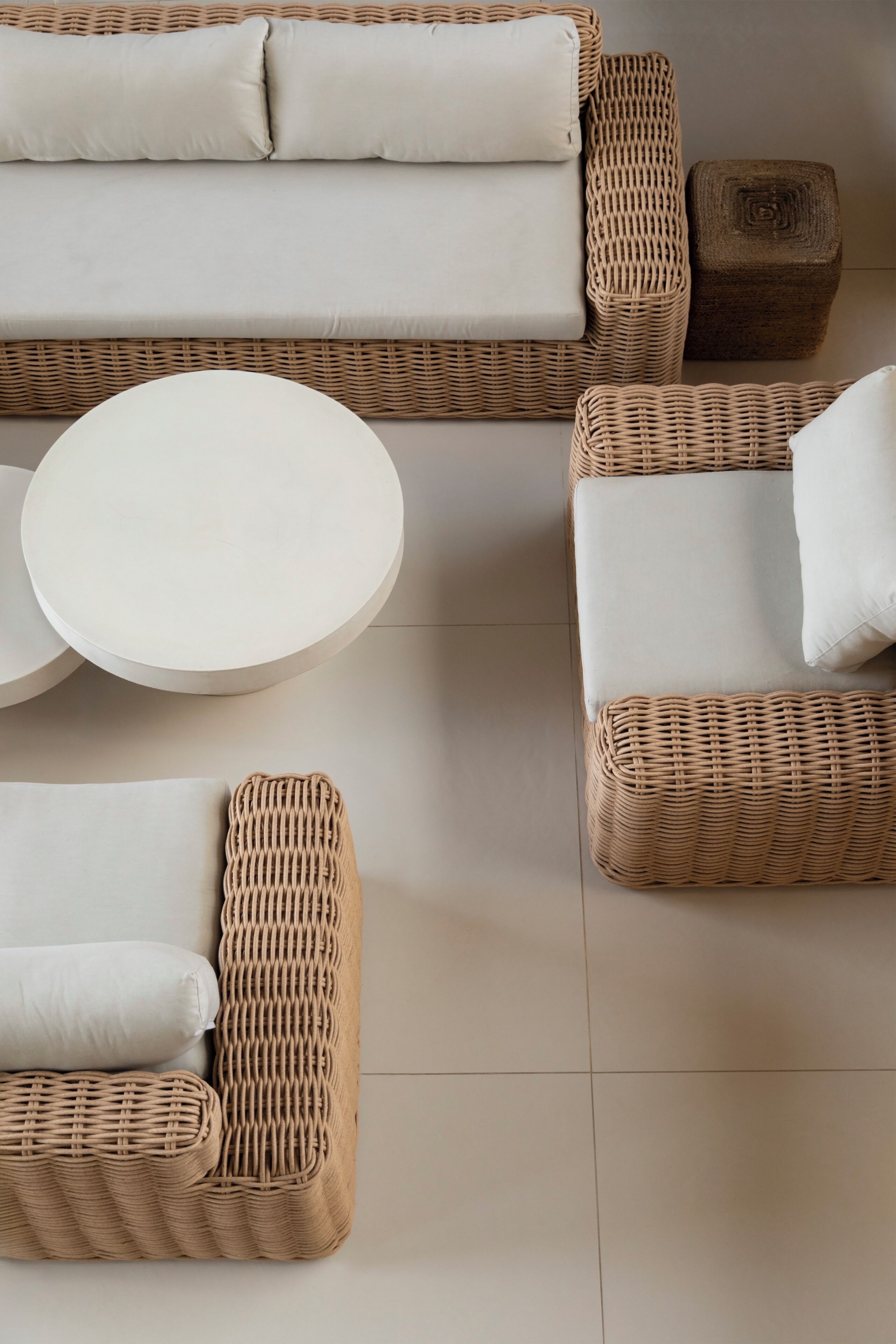 A wicker sofa with cushions, a round table, and a small wooden stool in a neutral-toned room.