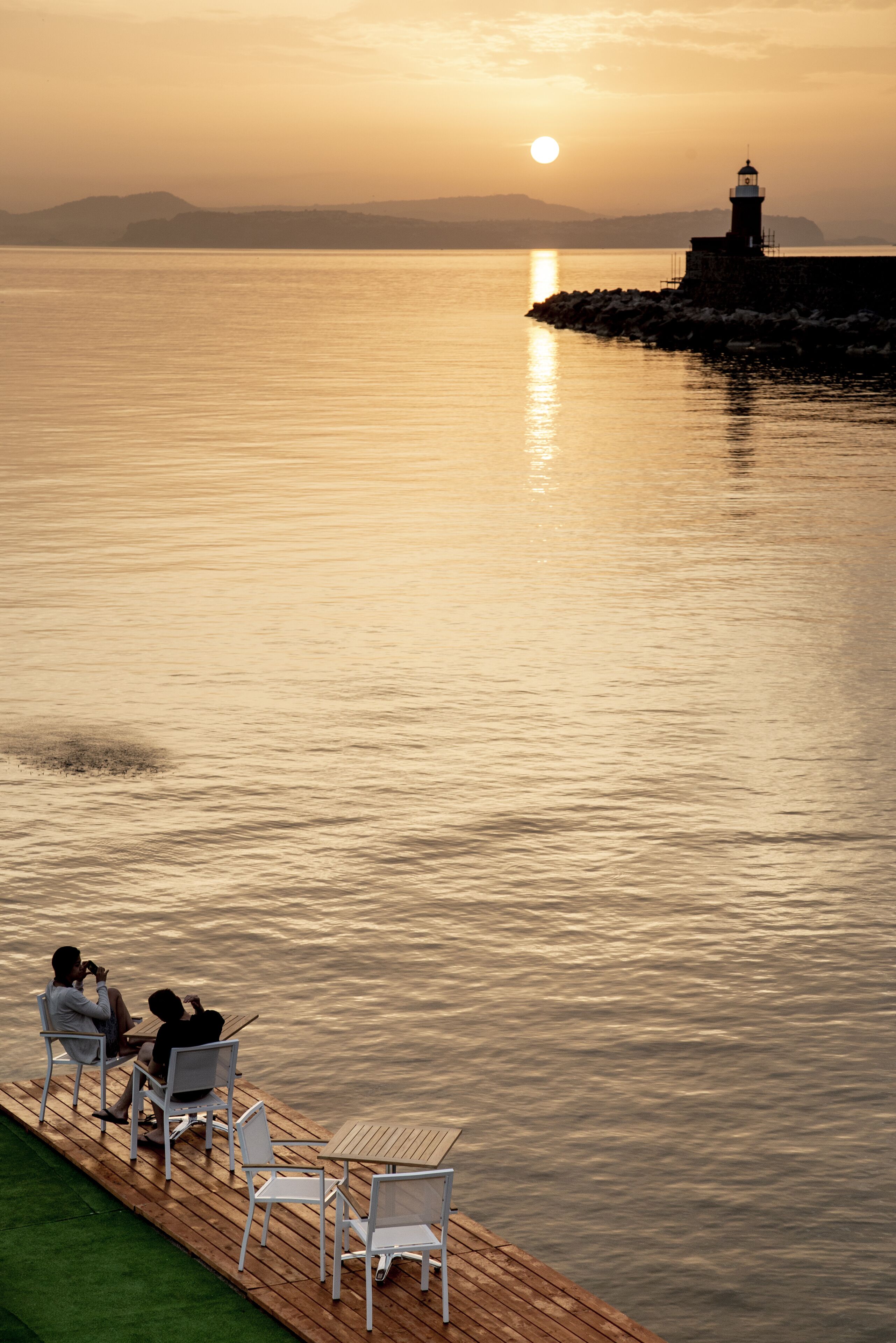 Une vue sur le coucher de soleil au-dessus d’une étendue d’eau calme, avec un phare au loin, une terrasse en bois avec des chaises et une zone herbeuse.
