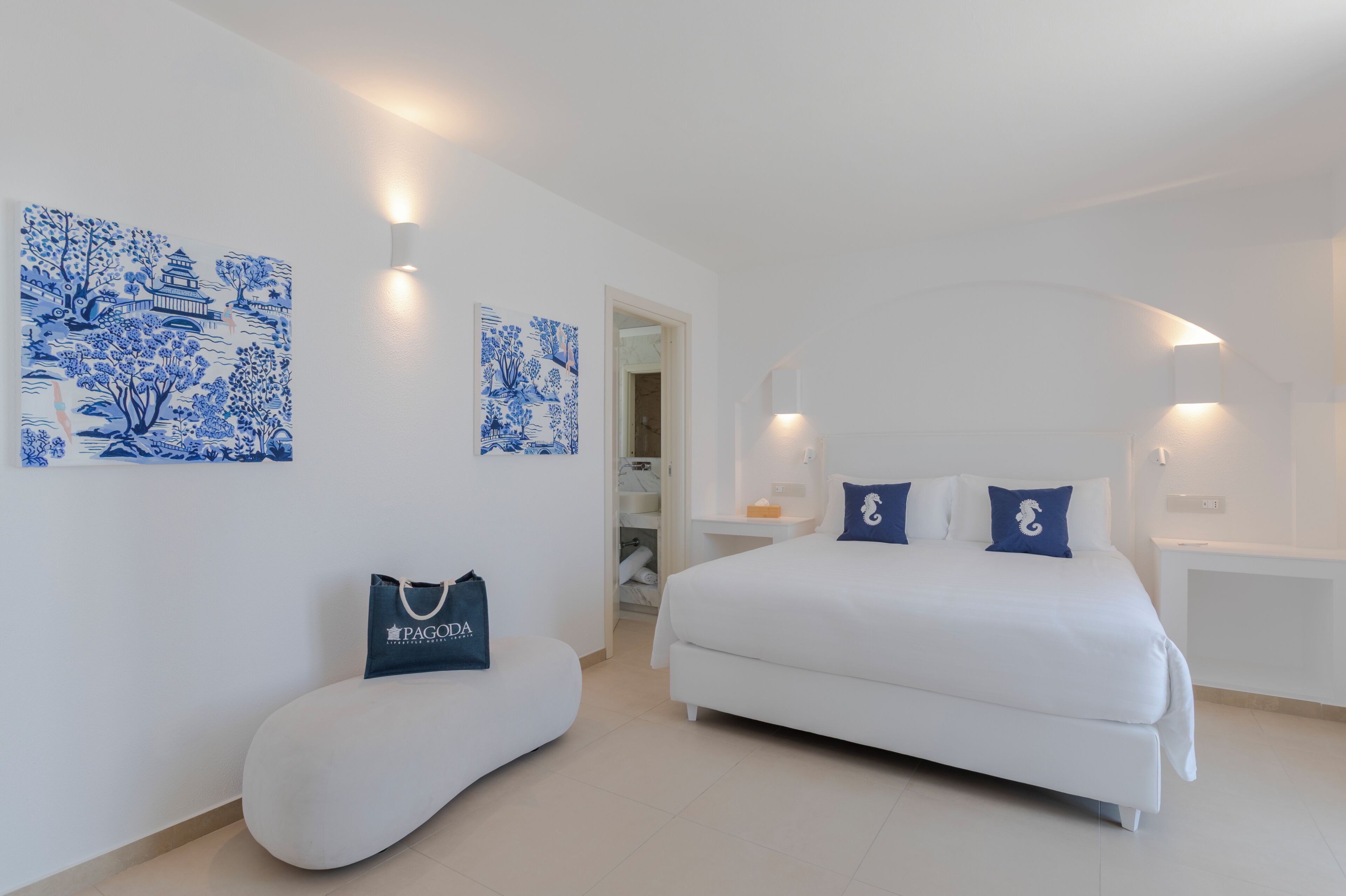 A modern hotel room with a white bed, two blue pillows, a white bench, and two blue wall art pieces.