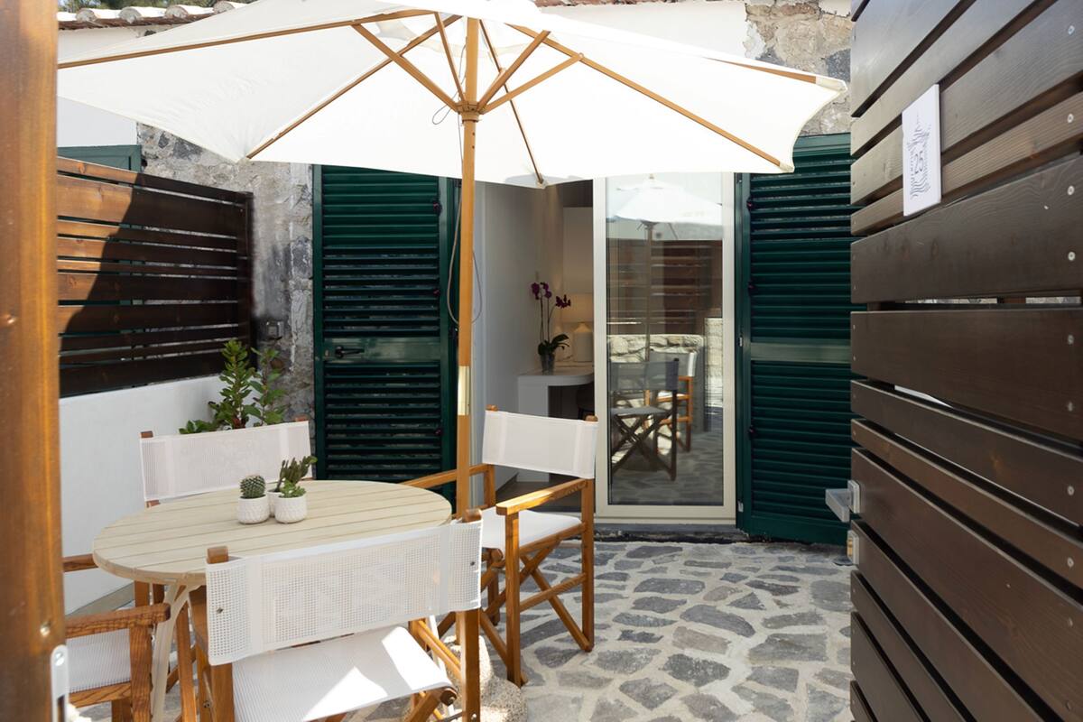 A patio with a wooden table and chairs, a white umbrella, and a stone floor.