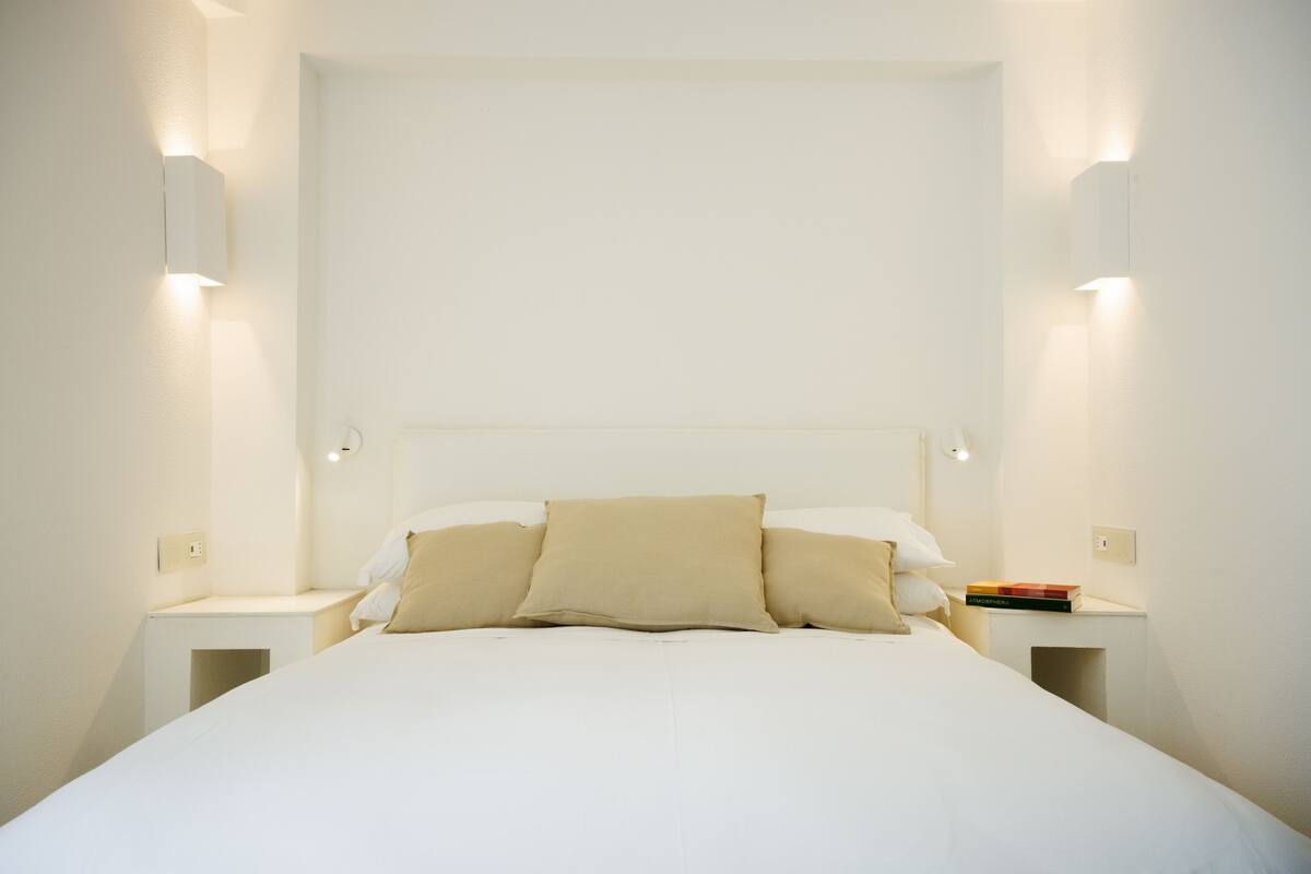 A neatly made bed with a white headboard, two bedside tables with books, and wall-mounted lights.