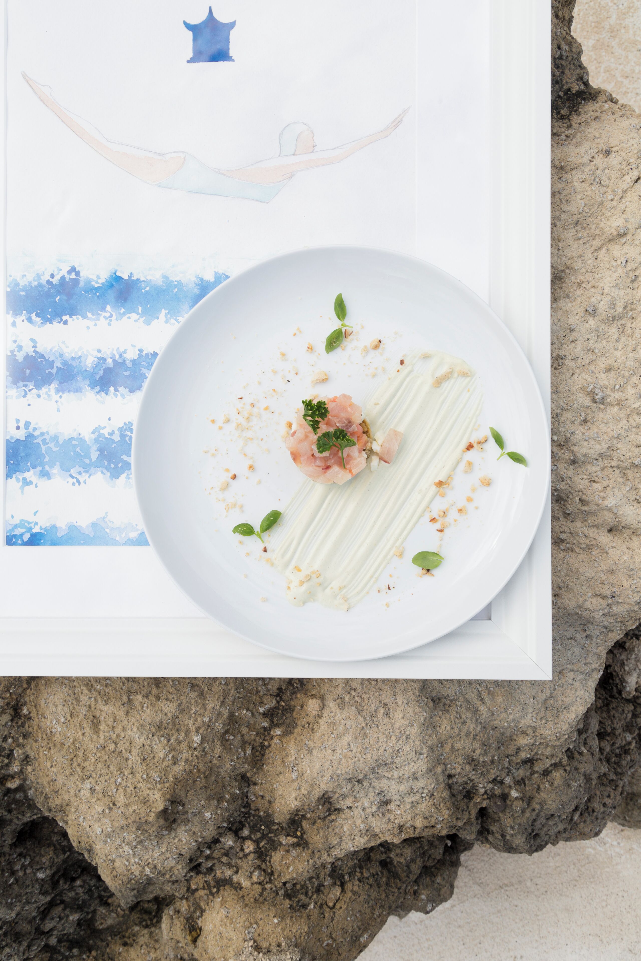 Ein weißer Teller mit einem weißen Dessert, verziert mit grünen Blättern und rosa Blüten, auf einem weißen Tisch.