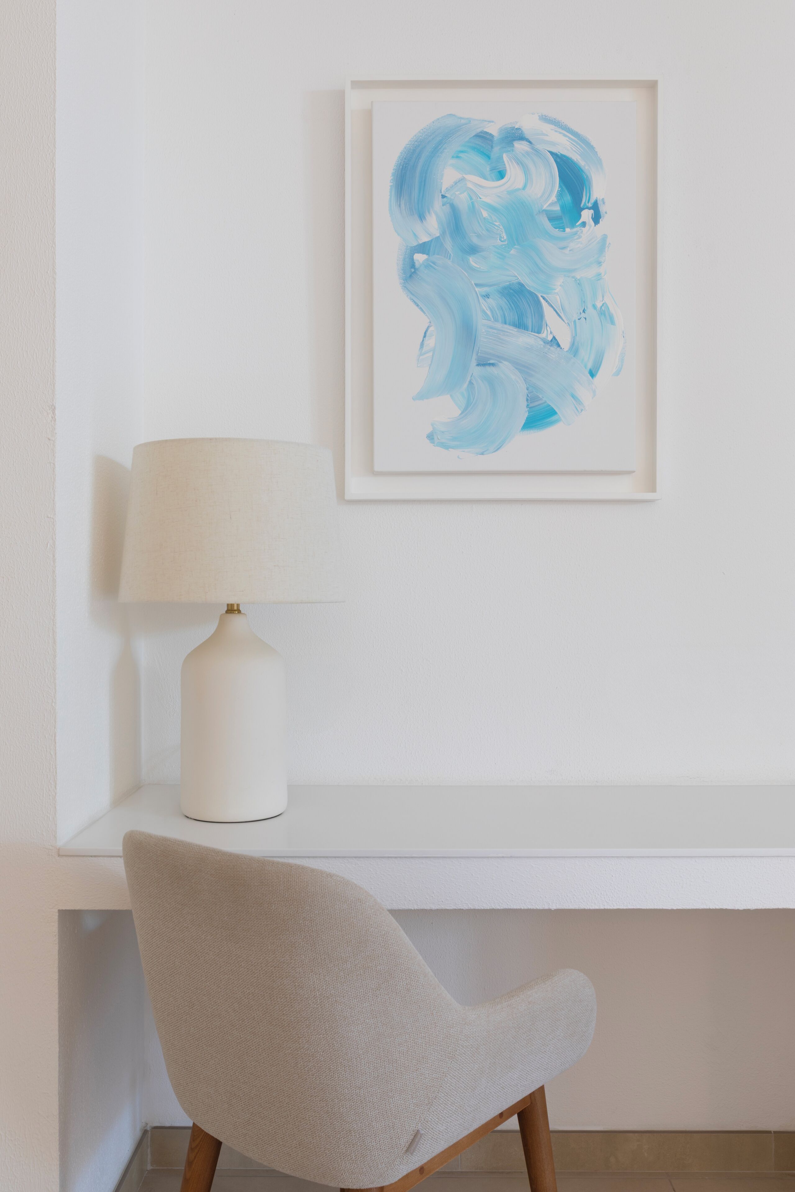 A white desk with a lamp and a chair, a framed abstract painting on the wall, and a white shelf below.