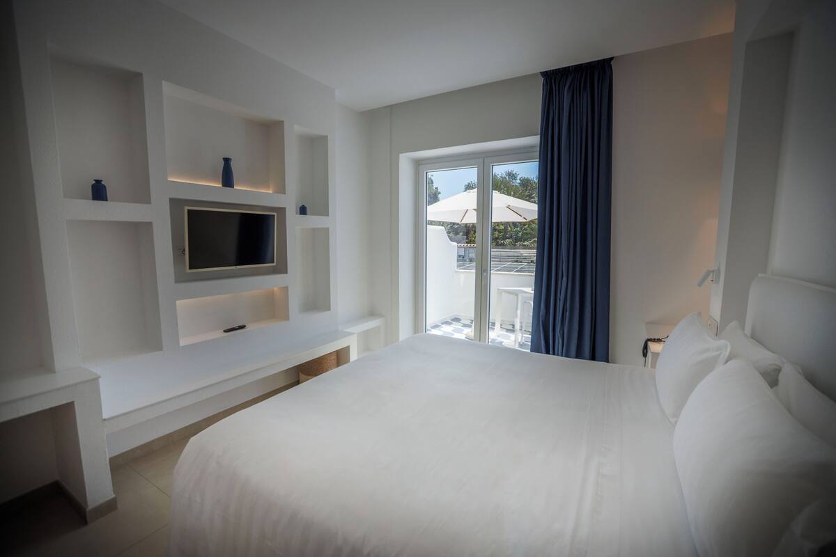 A hotel room with a large bed, a TV mounted on a white console, and a balcony with a view.