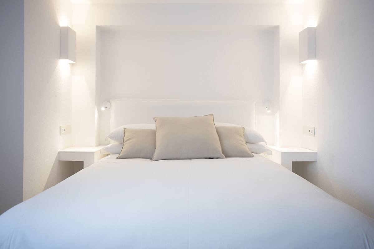 A modern, minimalist bedroom with a large bed, two bedside tables, and wall-mounted lighting.