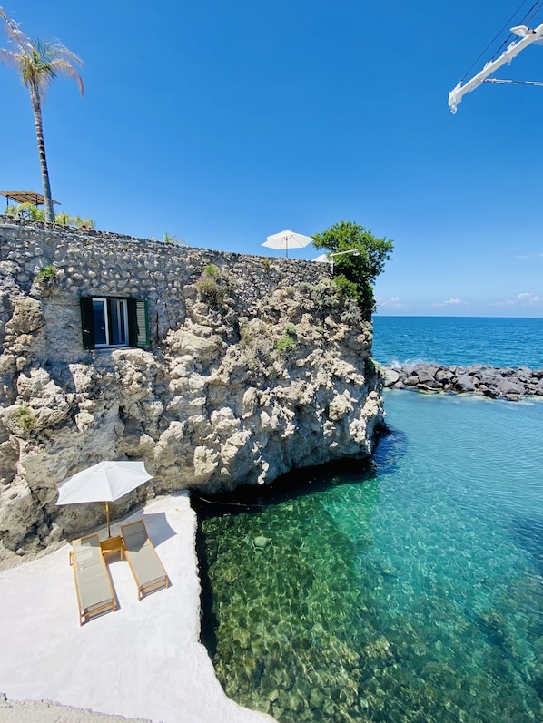Un edificio de piedra con una piscina cristalina, una palmera y vistas al mar.
