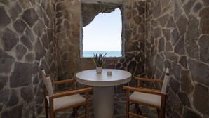 Una habitación con paredes de piedra, una mesa redonda y dos sillas, una planta en maceta y vistas al mar a través de una ventana.