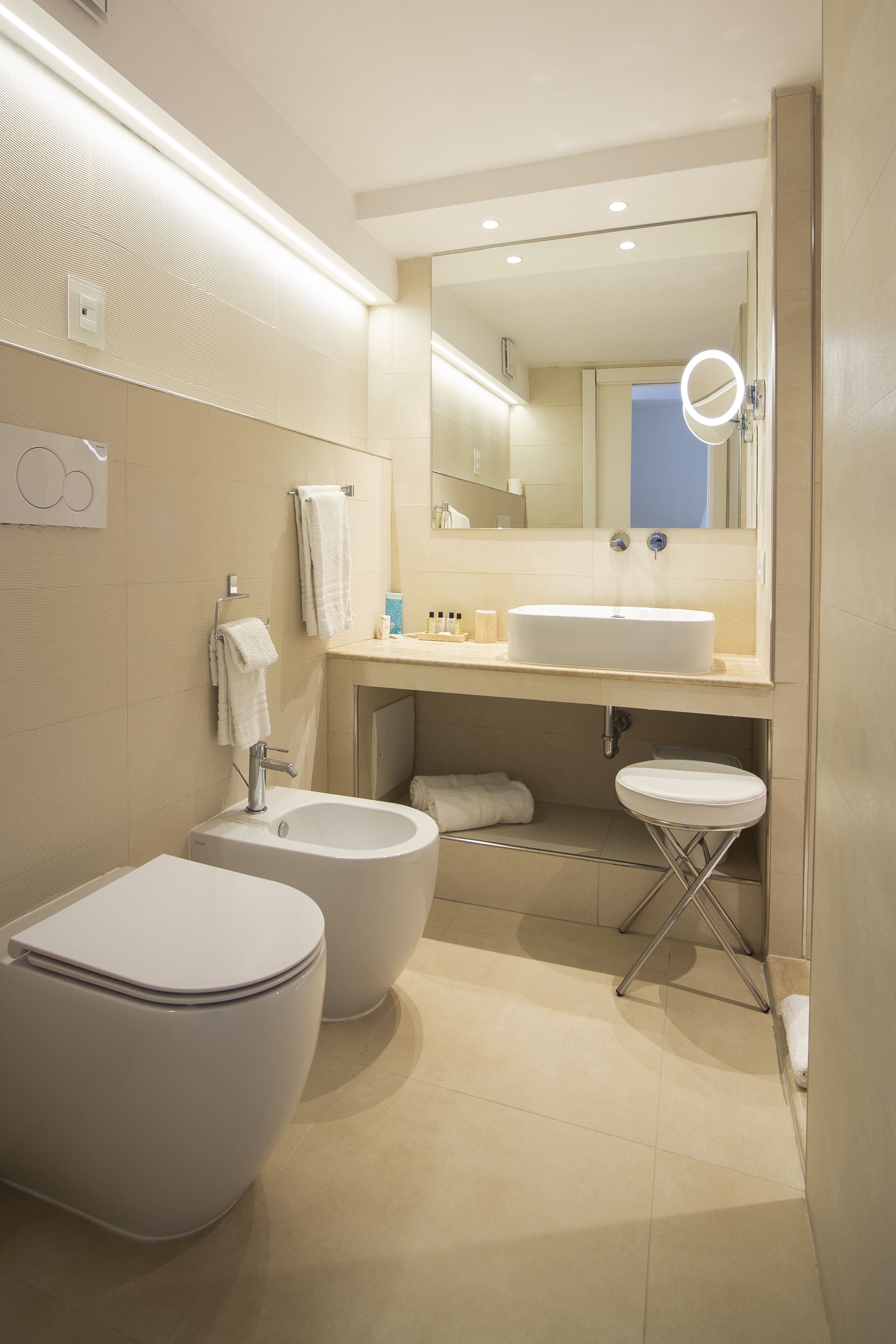 A modern bathroom with a large sink, two toilets, a mirror with a light, and a towel rack.