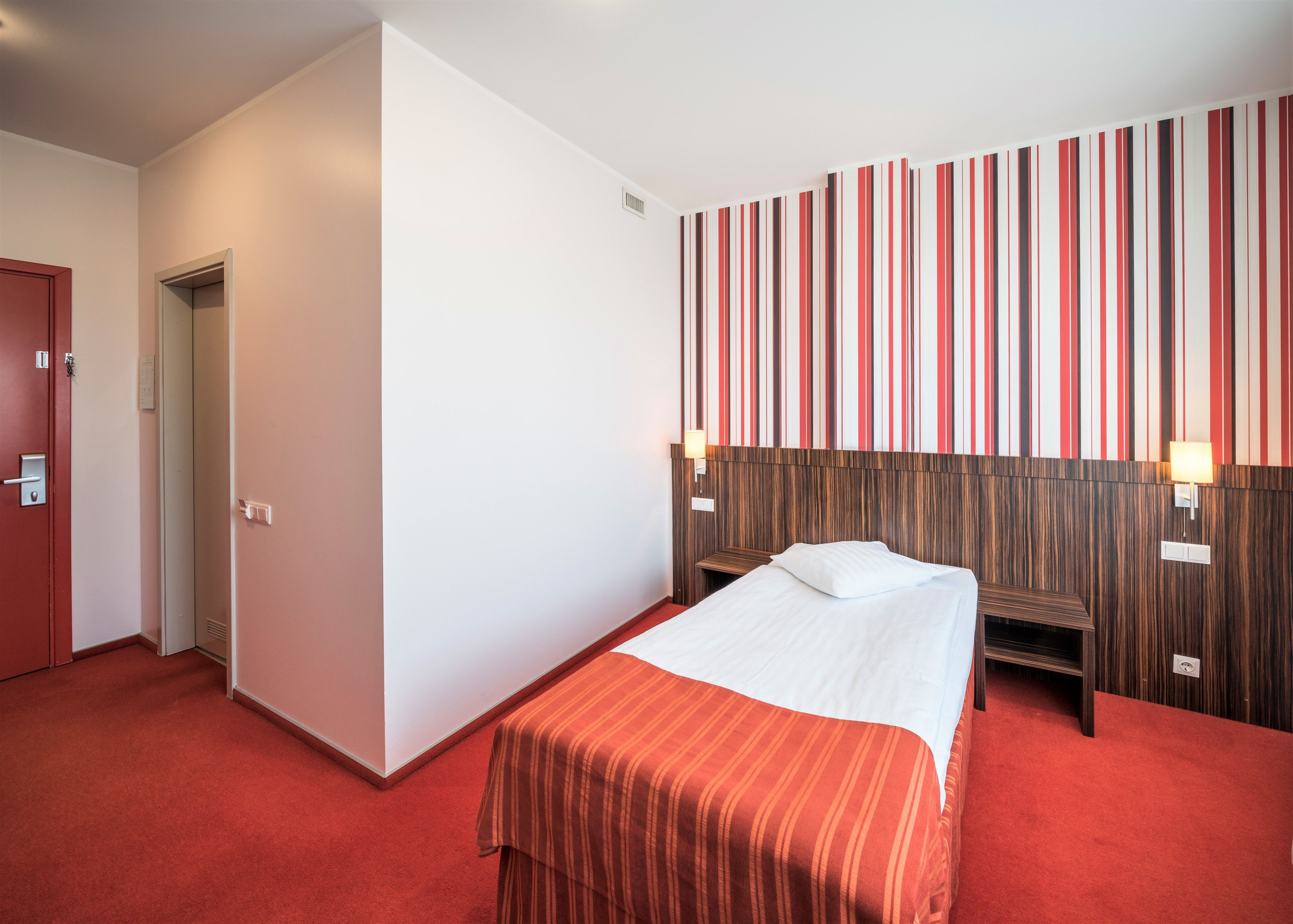 A hotel room with a bed, a wooden headboard, two bedside lamps, a red carpet, and a door.