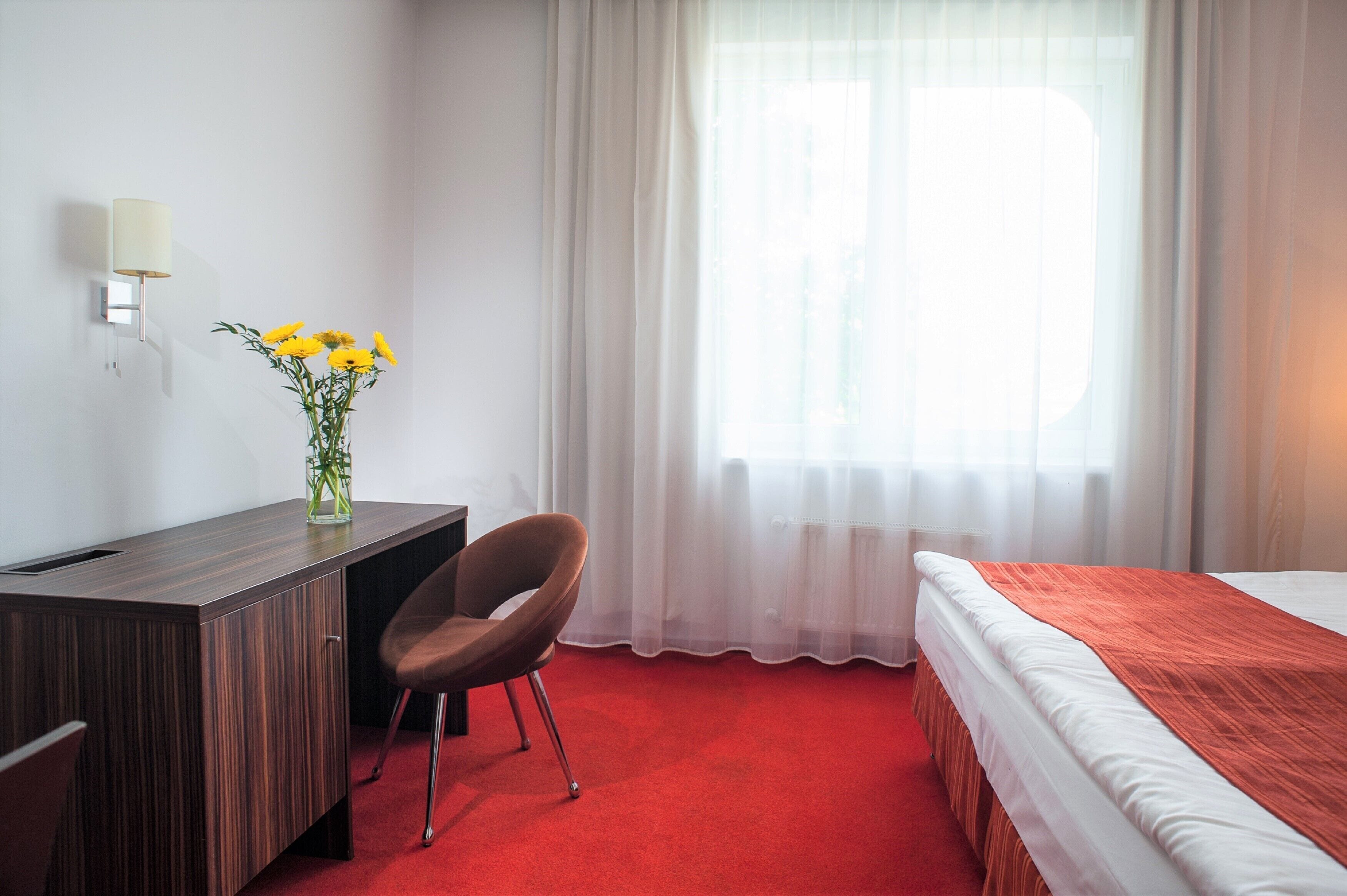 A hotel room with a wooden desk, a chair, a bed with a red blanket, a vase with yellow flowers, and a window with sheer curtains.