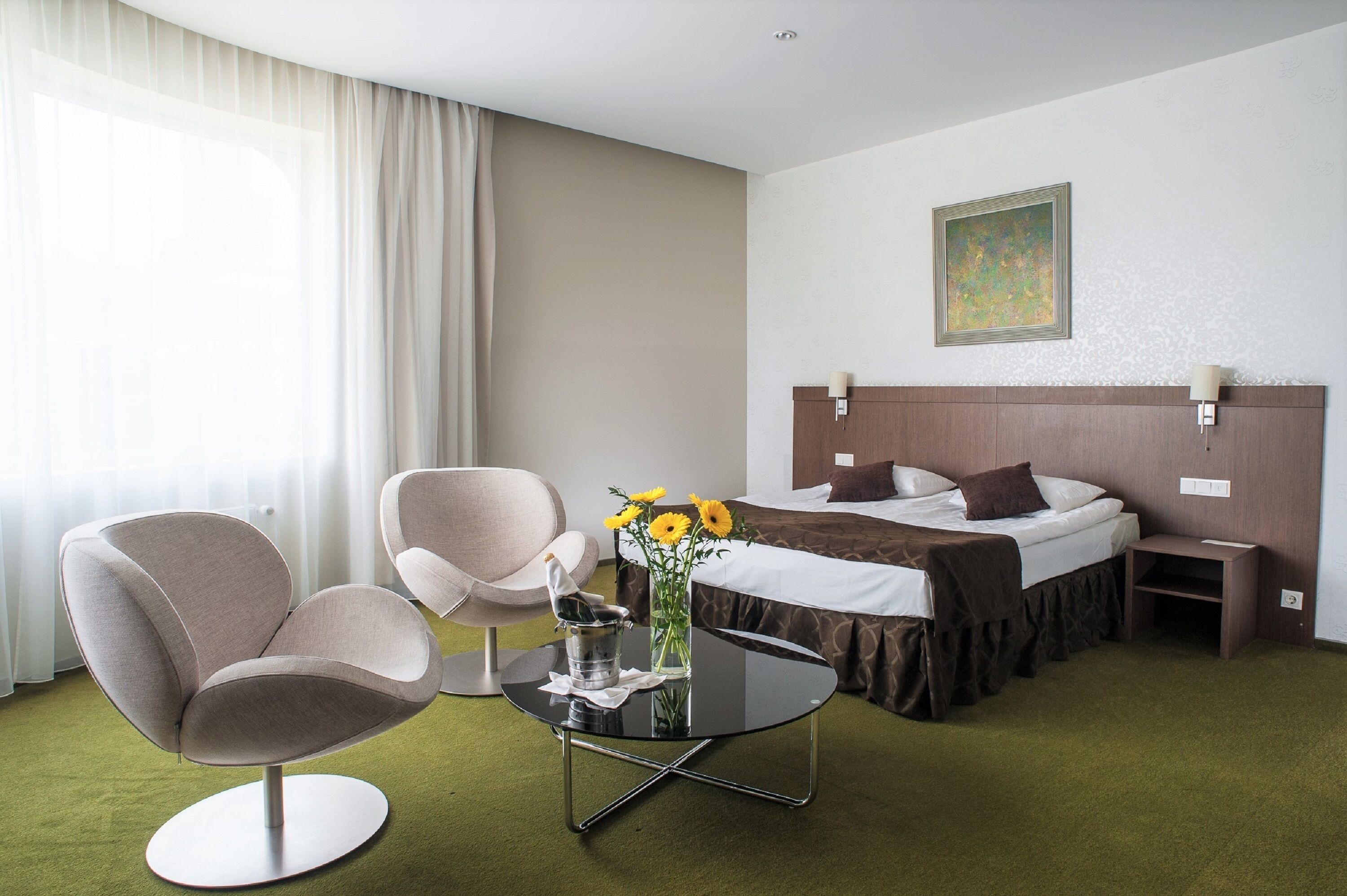 A hotel room with a bed, two chairs, a small table with a vase of flowers, and a painting on the wall.